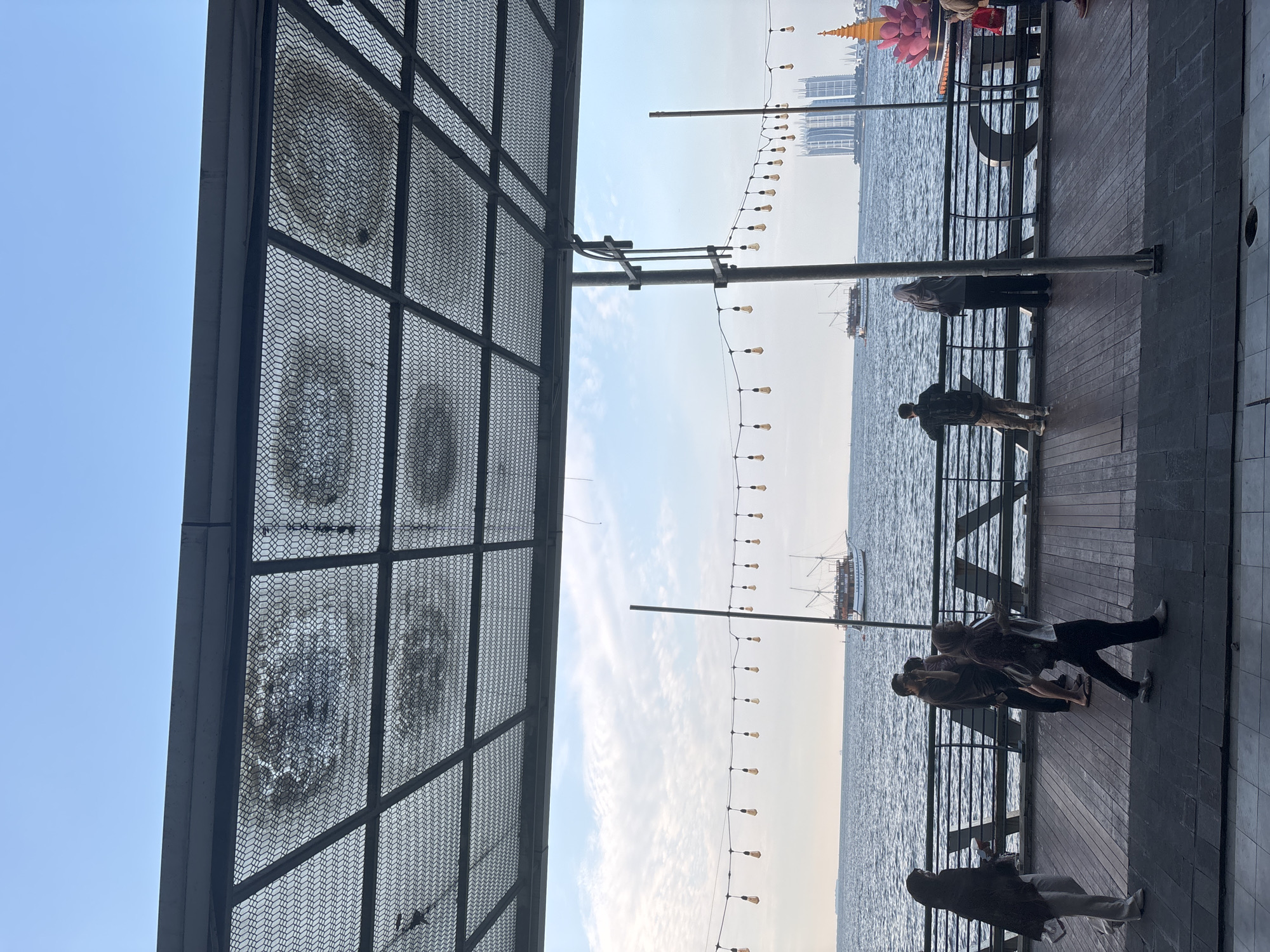 In North Jakarta, Indonesia, a scene unfolds on a waterfront promenade. The foreground is dominated by a dark wooden deck, partially obscured by a metal grate overhead, which casts a textured shadow. A string of incandescent bulbs hangs across the scene, adding a warm glow.
Several individuals are present on the promenade. To the left, a woman in a dark hijab and light pants walks away from the camera. Next to her, a group of three people stand, two facing the water. One of them, a man in a dark shirt, appears to be taking a photo with his phone. Further to the right, a woman in a light grey hijab stands with her back to the camera, looking out at the water. Another person, a man in a dark shirt and camouflage pants, stands to her left, also facing the sea. To the far right, a person in a red shirt is partially visible.
The middle ground features a dark metal railing that runs along the edge of the promenade. Beyond the railing, the dark blue water of the sea stretches out. Two boats are visible on the water: a larger, older-style wooden boat with sails and a smaller vessel further in the distance.
In the background, the city skyline of North Jakarta is visible. Two tall, modern white buildings with distinctive architectural features dominate the horizon. To the right of the buildings, a large, brightly colored pink lotus-shaped structure is illuminated. The sky above is a clear, pale blue with scattered white clouds. The overall impression is a lively urban waterfront setting.