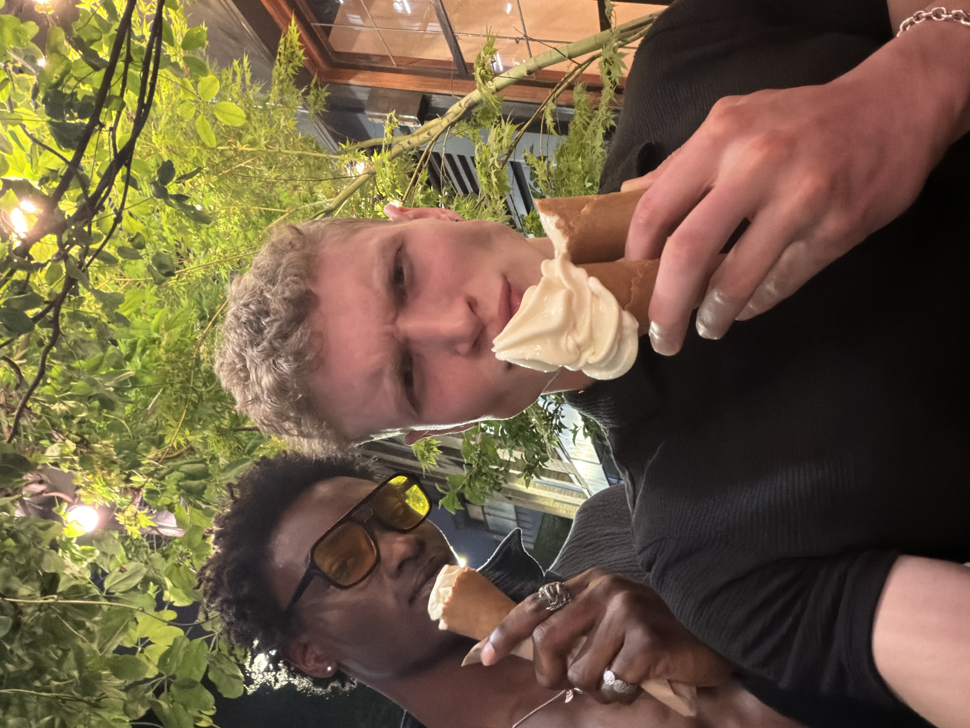 Two people pose with ice cream cones. The person on the left has dark skin, black hair, and is wearing yellow-tinted glasses and jewelry. The person on the right has fair skin, light hair, and holds two ice cream cones in one hand. Green foliage and artificial light are in the background. There are windows in the background as well.