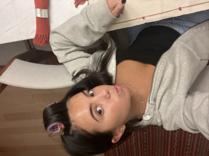 An overhead selfie captures a young woman seated at a table in an indoor residential setting in Lordelo do Ouro e Massarelos, Portugal. The woman, with dark hair, has a pink hair roller visible in a section of her hair on the right side of her head. She is wearing a light grey zippered hooded sweatshirt over a black strapless top and light blue and white striped bottoms. A delicate gold-toned necklace with small white beads is around her neck, and she has stud earrings in her left ear. Her lips are pursed into a "duck face" expression as she looks upwards towards the camera. Her left hand, with black-painted fingernails, rests on a white tablecloth adorned with a red striped pattern featuring small red heart-like motifs. On the white marble-patterned tabletop to her right, a rolled red textile item with fringed ends, secured by a silver ring, is present. Behind her, a dark red patterned textile, possibly a rug or wall hanging, is visible. To her right, a cream-colored upholstered chair with a dark wooden frame is partially visible, next to a light wood floor.