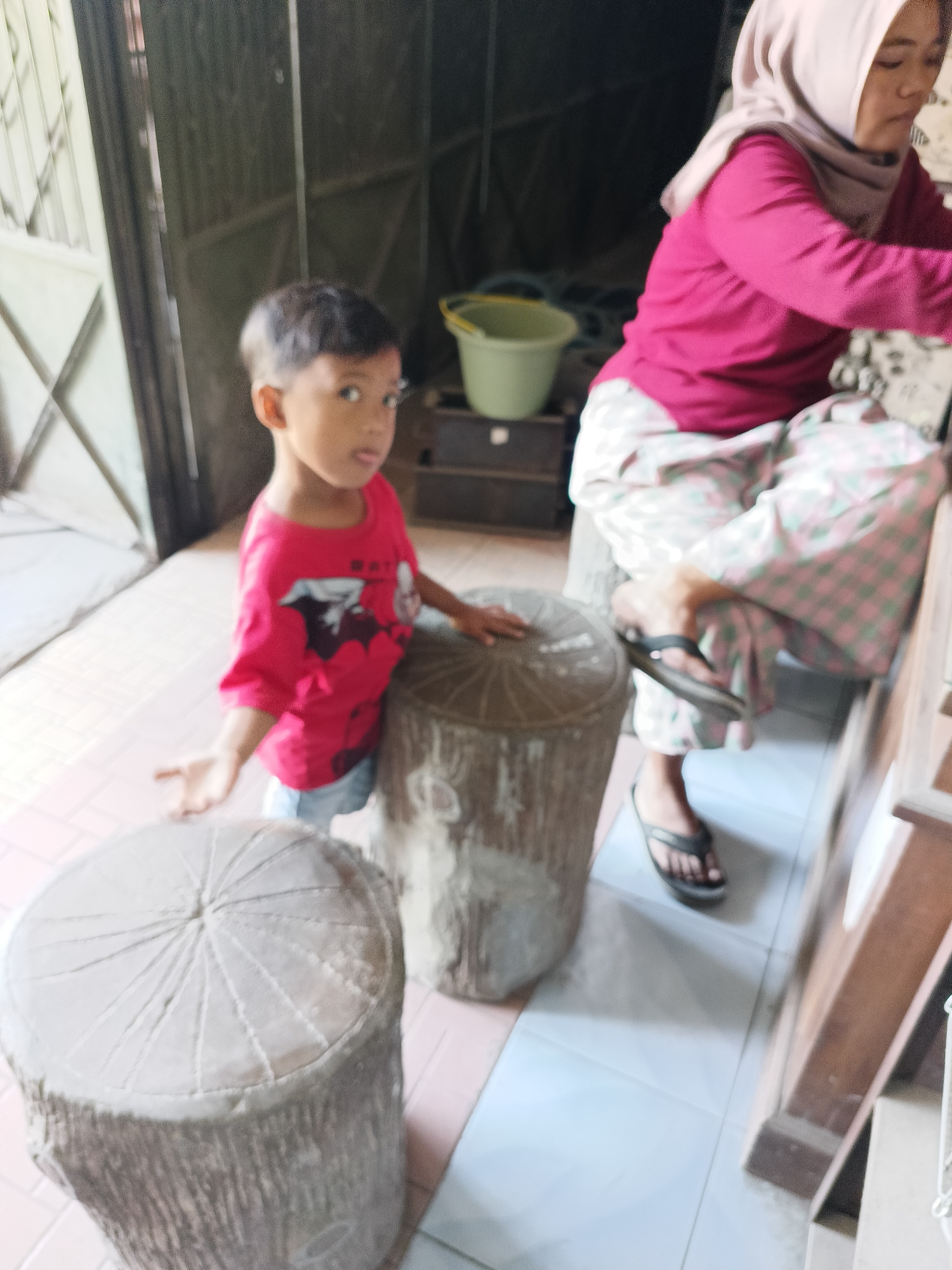 A young boy with dark hair and a red shirt stands near a wooden stool shaped like a tree stump. He is looking at the camera and has his tongue sticking out. A woman wearing a pink headscarf and a pink shirt is seated in the background, to the right of the boy. She is wearing patterned pants and flip-flops. The scene appears to be indoors, possibly in a home, in Sukorini, Indonesia. The floor is tiled, and there are other objects visible in the background, including a green bucket and what looks like a shelf unit.