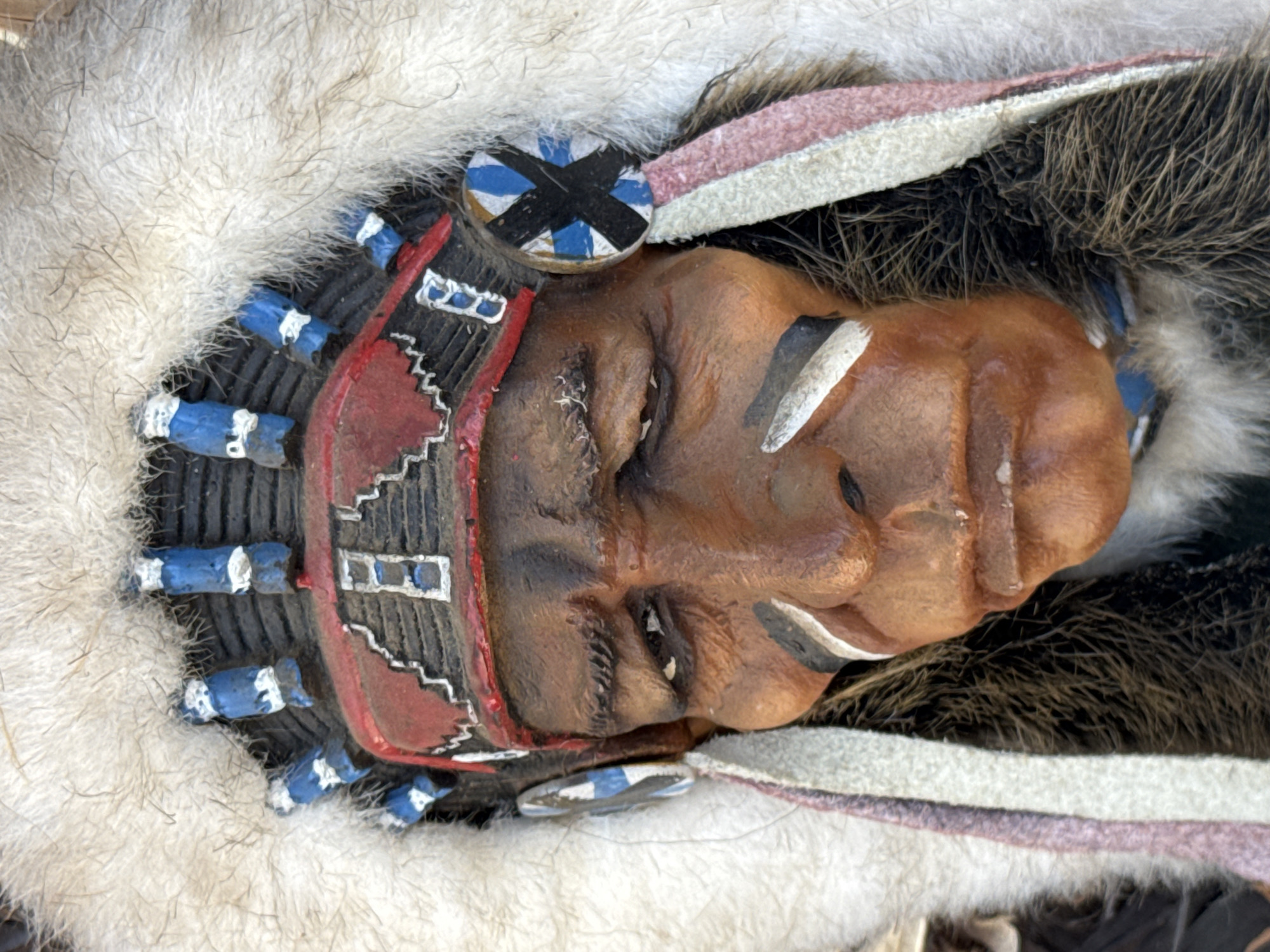 This is a close-up, head-and-shoulders, portrait of a wooden Native American headdress and bust. The headdress is adorned with blue and white feathers, red and black trim, and a fluffy white fur trim. The bust itself is carved to depict a Native American man with dark skin, dark hair, and facial war paint. The subject is wearing a beaded necklace and the bust is set against a dark, textured background. There are no discernible landmarks or environmental cues to suggest a specific location within Tel-Aviv, Israel.