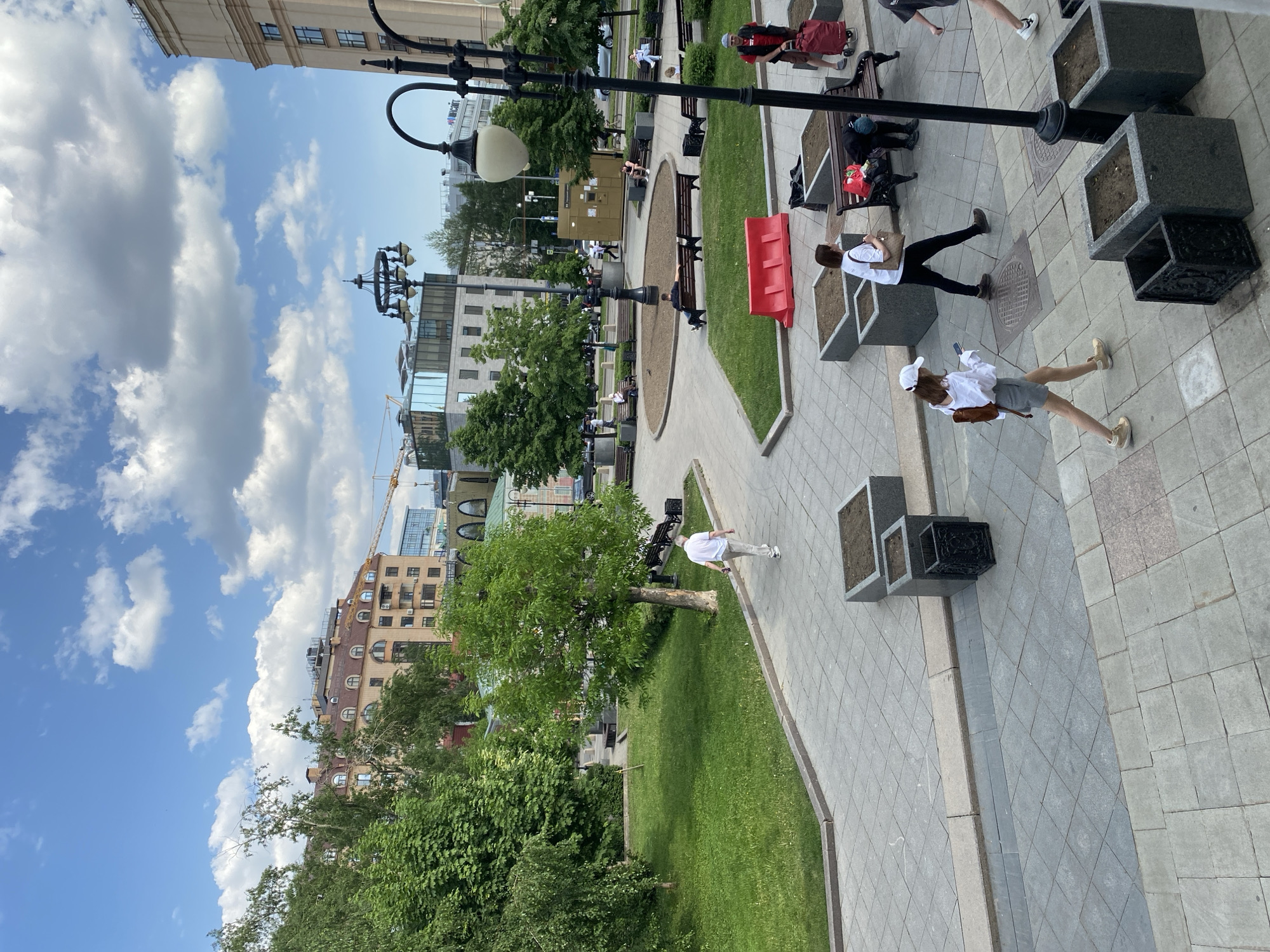 The image captures a view from above, looking down at a park in the city of Moscow, Russia. A brick walkway paved with tiles and planters extends through the park, dotted with benches, trees, and patches of grass. Several people are present, either walking or sitting on the benches. The architecture surrounding the park includes buildings of varying heights and styles. A construction crane is visible in the background. The sky is a clear blue with fluffy white clouds.