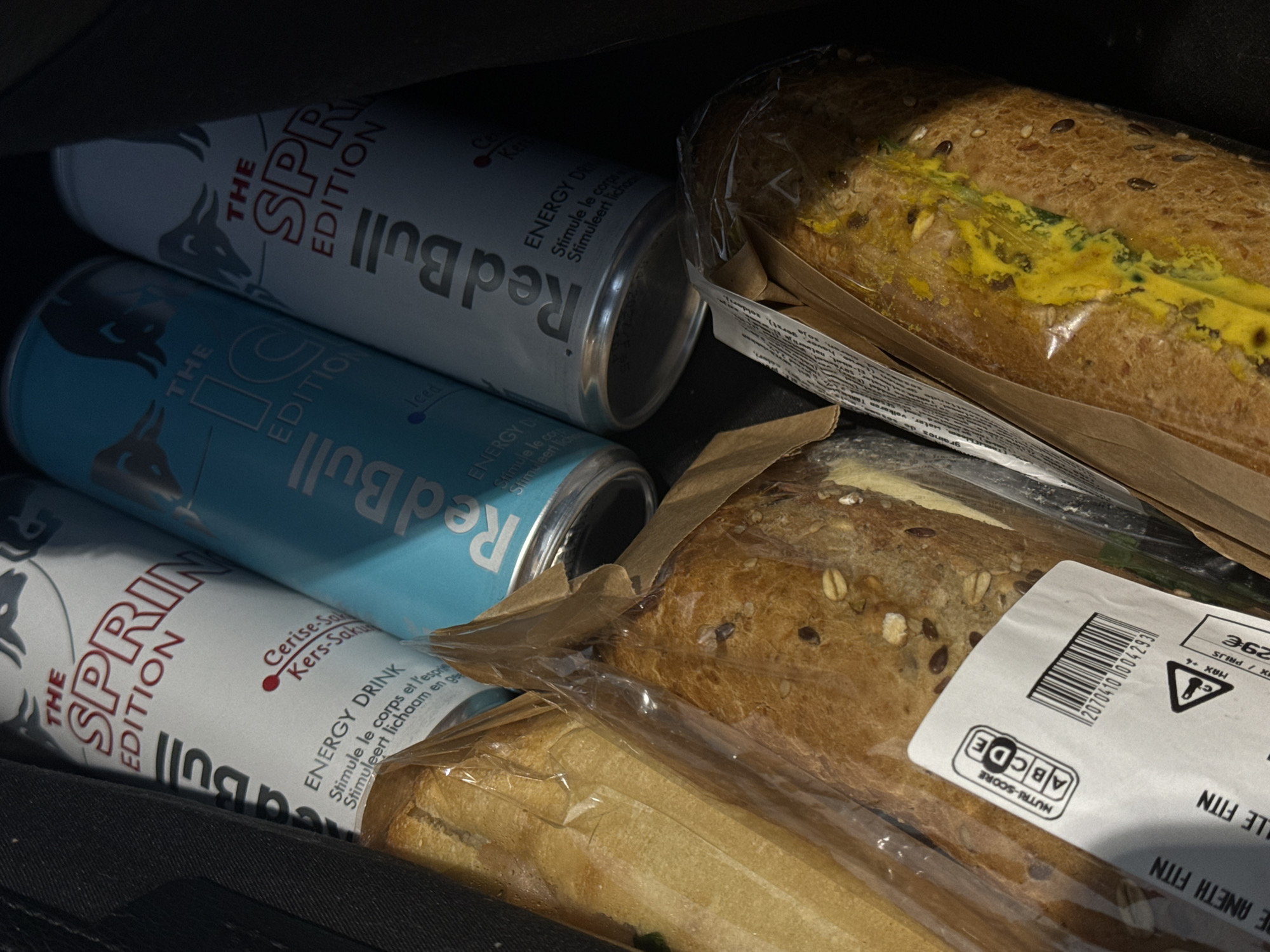The image shows a collection of items inside a dark bag. There are three Red Bull energy drink cans: two "The Spring Edition" cans with a cherry-sakura flavor, and one "The Iced Edition" can. Two packaged sandwiches are also present. The labels on the sandwiches include a barcode "2070410004293" and a nutrition score with the letter grade "C". Based on the text "E ANETH FITN LE FITN" visible on one of the sandwich labels, it's likely the sandwiches were purchased at a "Ftin" store, a local chain in Luxembourg. Considering the prevalence of this store chain in Esch-sur-Alzette, Luxembourg, it's plausible the items were purchased or are located there.