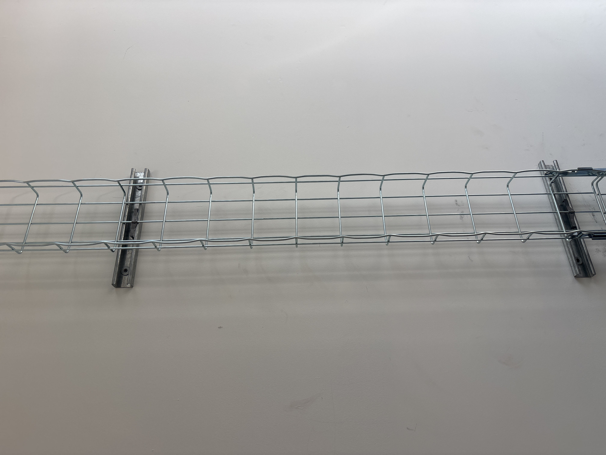 A wire cable tray is mounted horizontally on a light gray wall. The tray is constructed of galvanized metal and has a grid pattern. It is secured to the wall by two vertical supports. The location is unknown and not discernible from the image.
