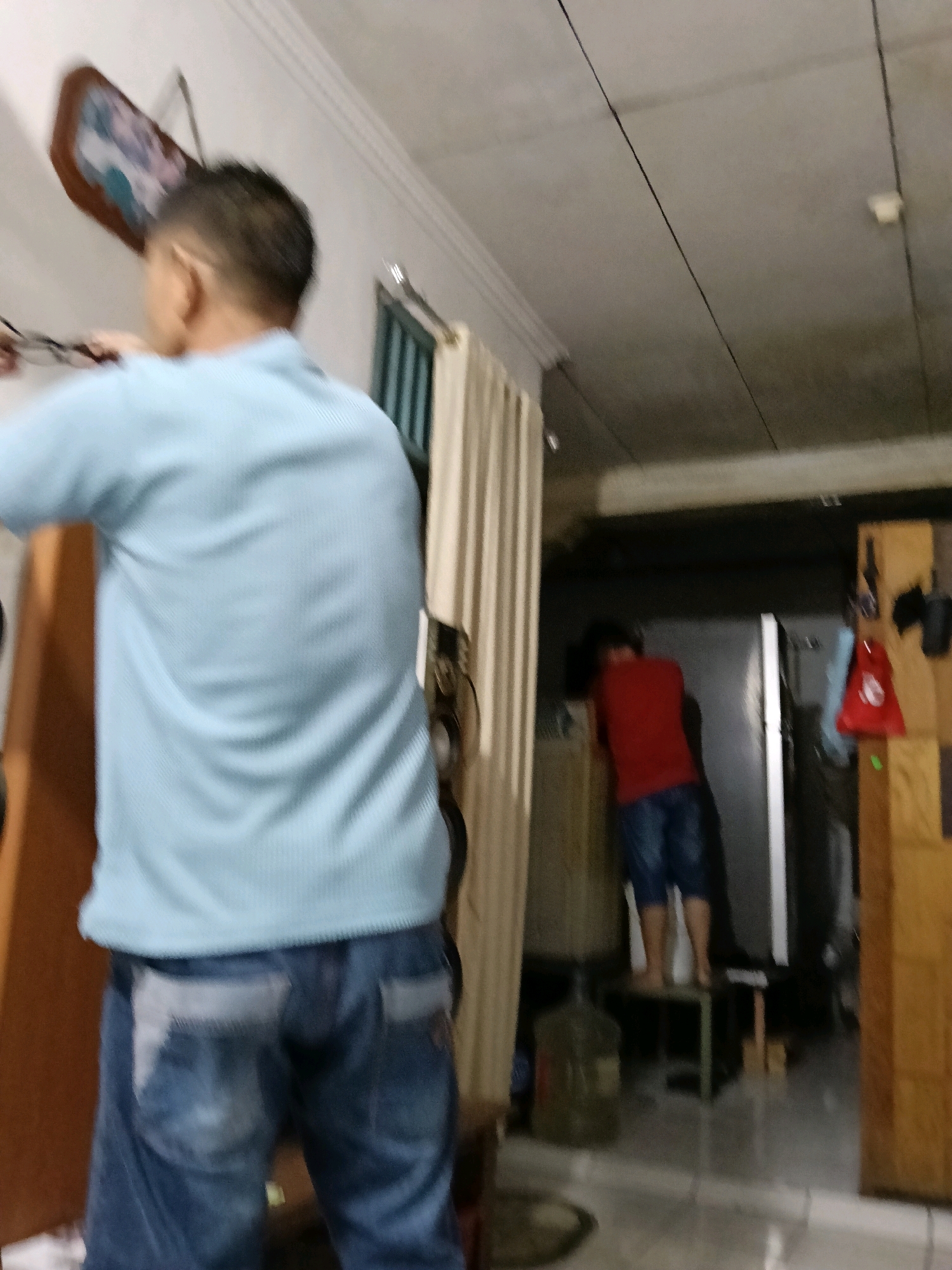 The image depicts an indoor scene, possibly a residential interior, in Jatimulya, Indonesia. In the foreground, a person wearing a light blue t-shirt and blue jeans is partially visible from the back, facing away from the camera. The person appears to be holding a pair of glasses in their left hand. To the right of the foreground figure, further in the background, another person wearing a red t-shirt and blue shorts is standing on a dark-colored stool. This second person is leaning forward, with their head down, near what appears to be a refrigerator. The environment includes a light-colored ceiling with visible electrical wires, a beige-colored vertical partition or door on the left, and wooden shelving units on the right side of the frame. Water cooler jugs are visible on the floor. The lighting is dim, creating a low-light environment.