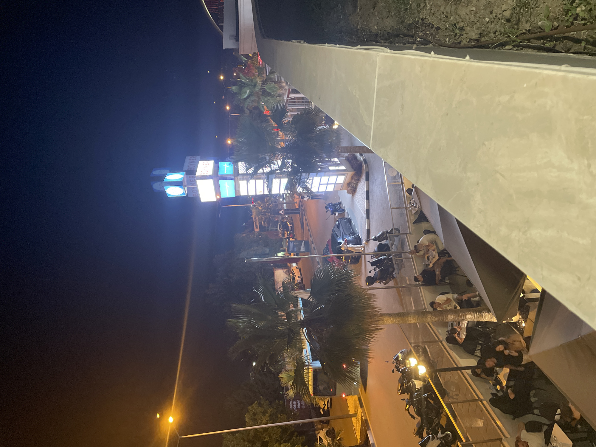 This is a night-time, high-angle shot of a street scene in Yunus Emre, Turkey. A tall, illuminated signpost with multiple screens displays the word "BOYNER" multiple times and abstract graphics. Several palm trees are present. In the foreground, a light-colored concrete wall runs along the bottom right of the frame. Below this wall, on a lower level, a group of people are seated at tables, some under umbrellas. To the left, a street is visible with parked motorcycles and cars. A bus is also seen driving on the street. The sky is dark, indicating it is nighttime, with streetlights and the illuminated sign providing the primary light sources.