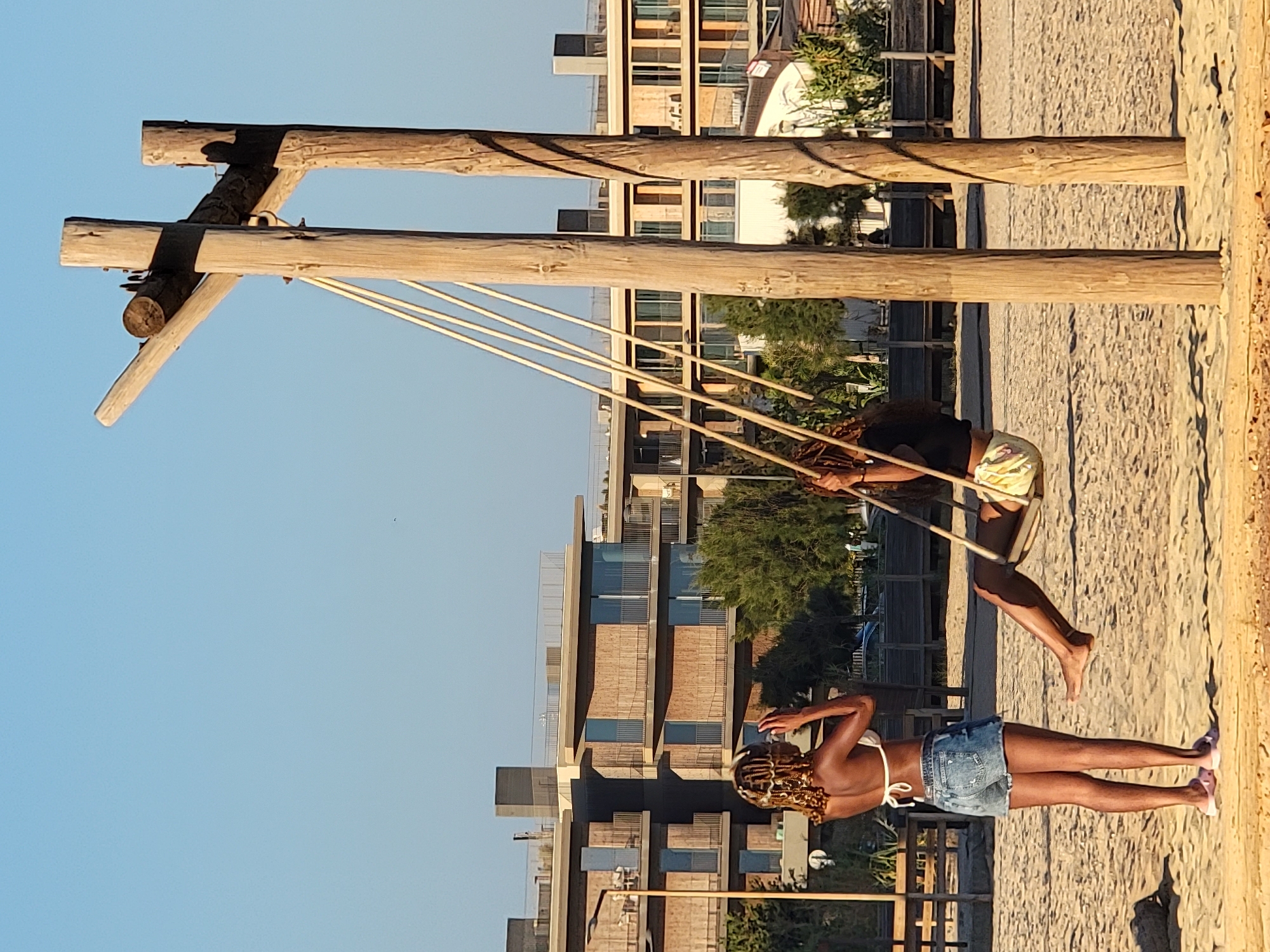 On a sandy beach, a wooden swing set with two vertical posts and a horizontal beam supports a swing with two ropes. A woman with braided hair, wearing a white bikini top and denim shorts, stands on the left, looking towards the right and touching her hair. A second woman, also with braided hair, is seated on the swing, wearing a black top and patterned shorts, with her legs extended. In the background, a modern multi-story building with wooden accents and balconies is visible, along with trees and a clear blue sky. The scene appears to be on the beachfront in Quarteira, Portugal.