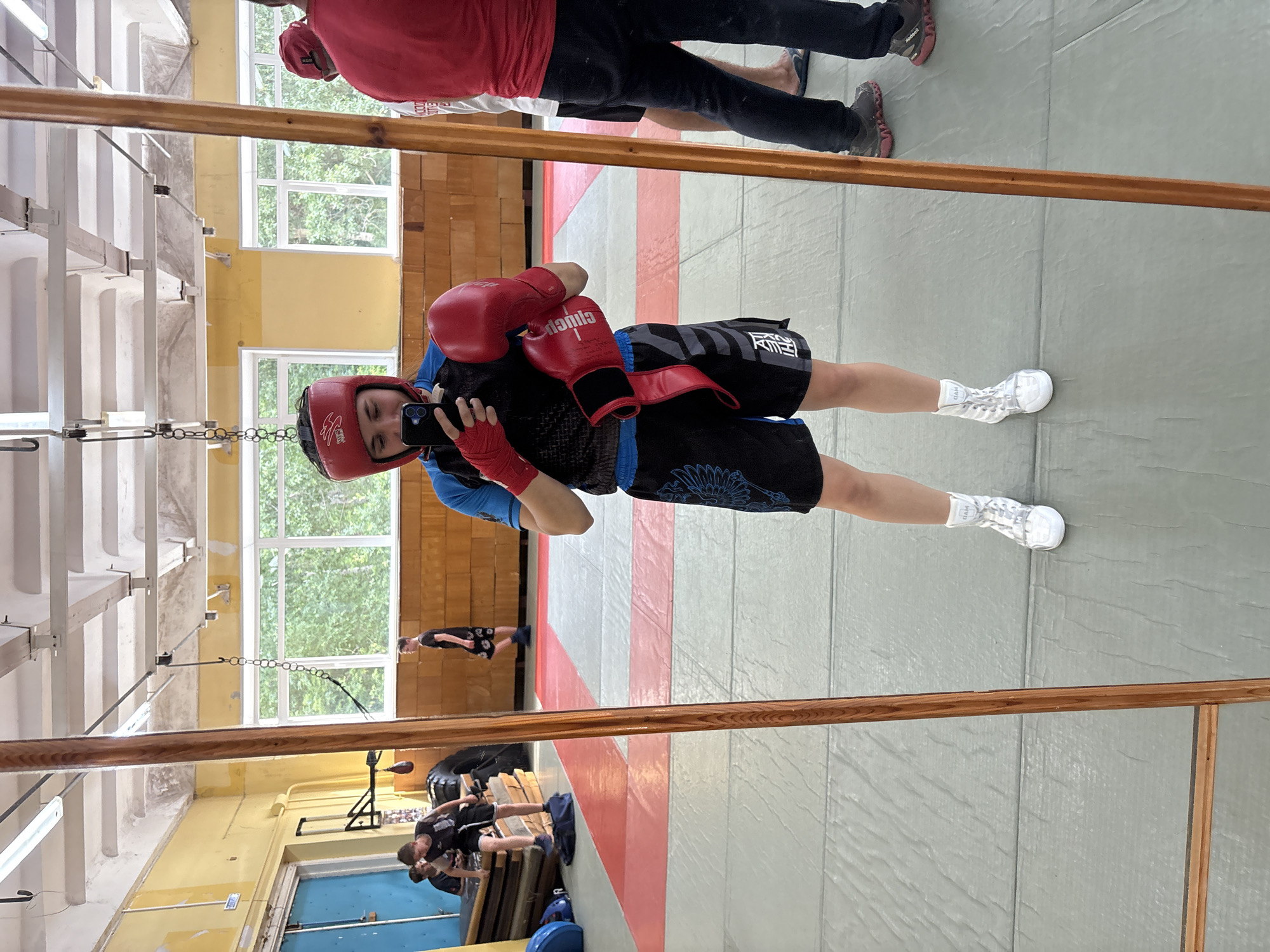 The image is a full-length mirror selfie taken in what appears to be a gym or dojo in Chaikovsky, Russia. The primary subject is a young person wearing a red boxing helmet, red boxing gloves, blue and black athletic shorts with a tribal-like design, and white high-top athletic shoes. They are holding a smartphone to take the picture.
In the background, visible through the mirror's reflection, are several other individuals, also dressed in athletic attire. Two young males are positioned near a stack of mats. Another person, wearing a red shirt and dark pants, stands to the right. The environment features wooden paneling on the walls, large windows looking out onto trees, and fluorescent lighting fixtures on the ceiling. The floor is covered with a mat, marked with red and grey sections. A wooden pillar, part of the mirror's frame or a structural element, is prominent in the foreground. The overall impression is a training session in progress or about to begin.