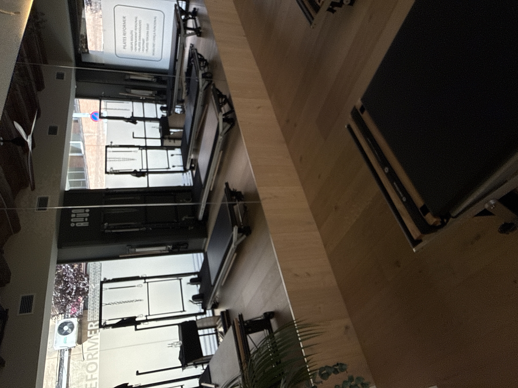 The image depicts the interior of a Pilates studio, likely located in Barcelona, Spain, based on the visible streetscape. Several Pilates reformer machines are arranged in rows, facing large windows that offer a view of the exterior. No people are visible in the immediate scene. The overall environment appears clean and well-lit, indicative of a professional fitness facility. A sign with the word "REFORMER" is also visible.