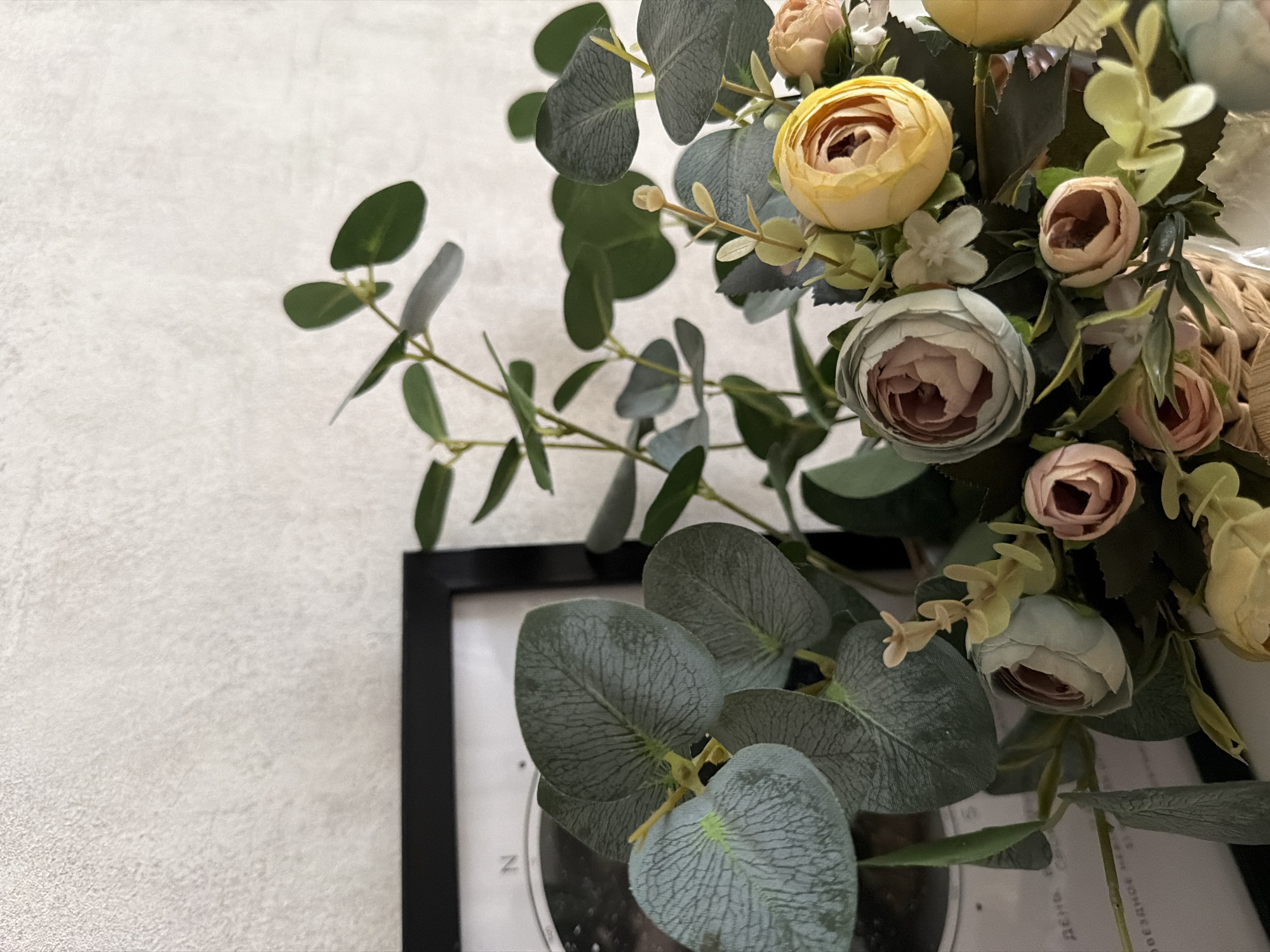 The image shows a close-up view of a floral arrangement in a wicker basket. The arrangement features a variety of artificial flowers in shades of pink, yellow, and light blue, interspersed with green eucalyptus leaves and stems. A black-framed picture is partially visible on the left side of the image, behind the foliage. The background is a textured, off-white wall. The lighting suggests an indoor setting. There are no people visible in the image. The location is specified as Semiluki, Russia, however, there are no visual cues in the image that confirm or indicate this specific location.