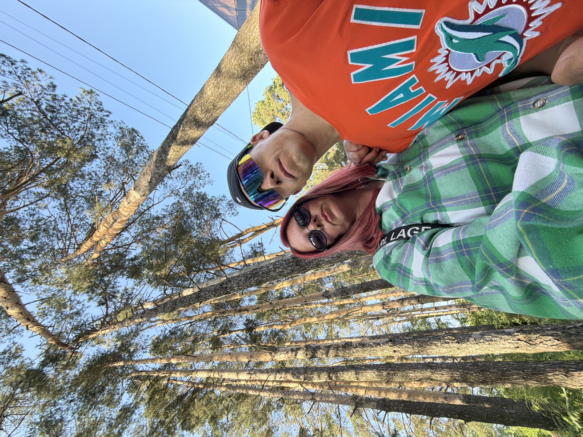 The image shows two individuals in an outdoor setting dominated by tall pine trees. The perspective is from a low angle, looking upwards. A person wearing an orange shirt with "MIAMI" lettering and a Miami Dolphins logo is seen above a person with pink hair, wearing sunglasses and a green and white plaid shirt. The trees are the predominant background element, with their trunks and branches filling the frame. The blue sky is visible through the trees. The location is Chaikovsky, Russia.