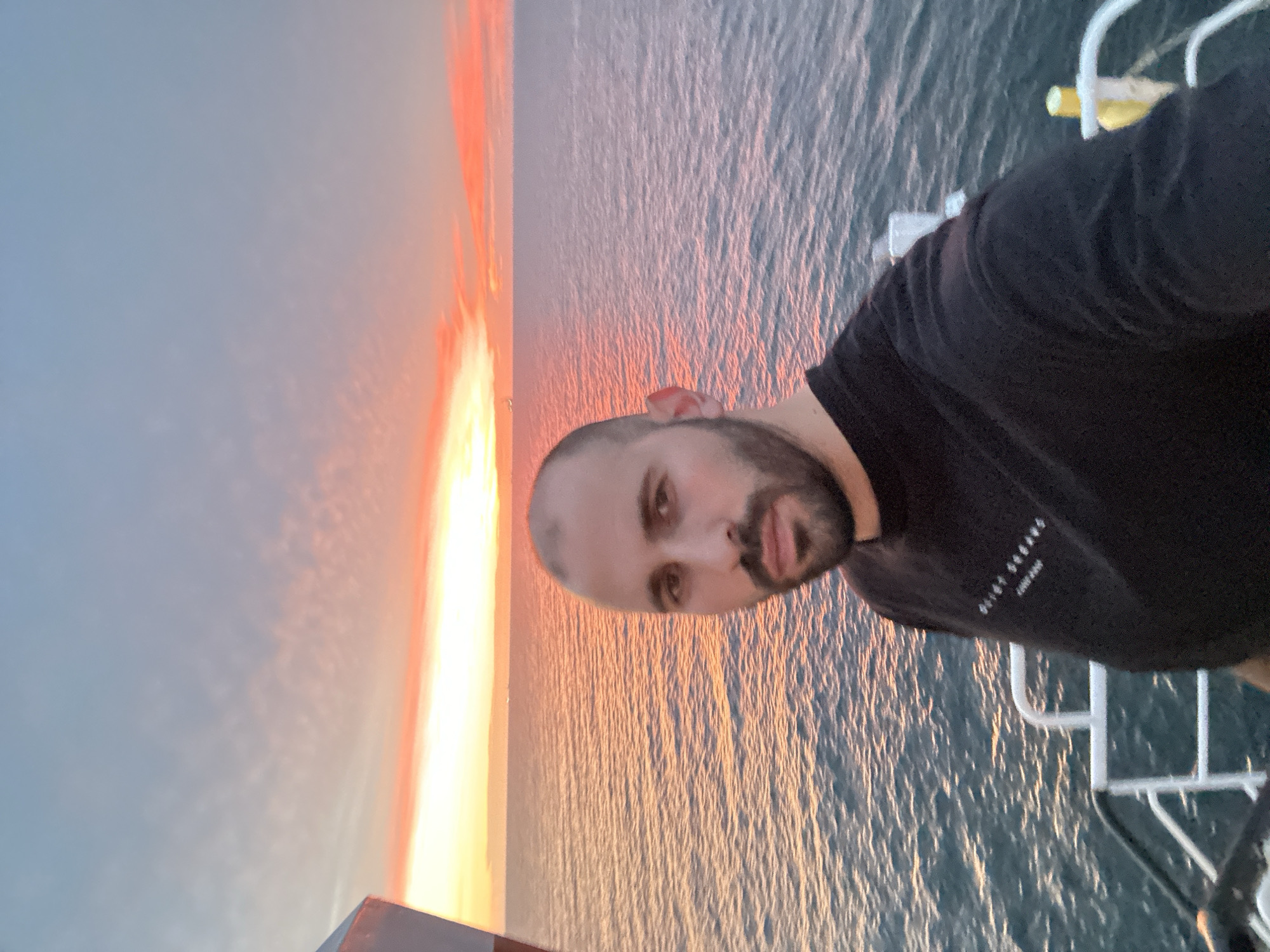 A bald man with a beard, wearing a dark long-sleeved shirt with white text, is centered in a close-up, chest-up shot on the deck of a vessel. He looks directly into the camera with a neutral expression. The background features a vibrant sunset over a calm sea, with the horizon line visible. The sky transitions from bright orange and red hues near the horizon to softer pinks and light blue at the top, showing subtle cloud patterns. The ocean surface reflects the sunset colors, appearing orange and pink near the horizon and a darker blue-green closer to the vessel. Parts of the vessel's white railing are visible in the lower right, and a dark structure is partially visible on the far left. The man's shirt text reads "QUIET STREAMS" and "LONDON". The location is at sea, with no discernible city or land features.