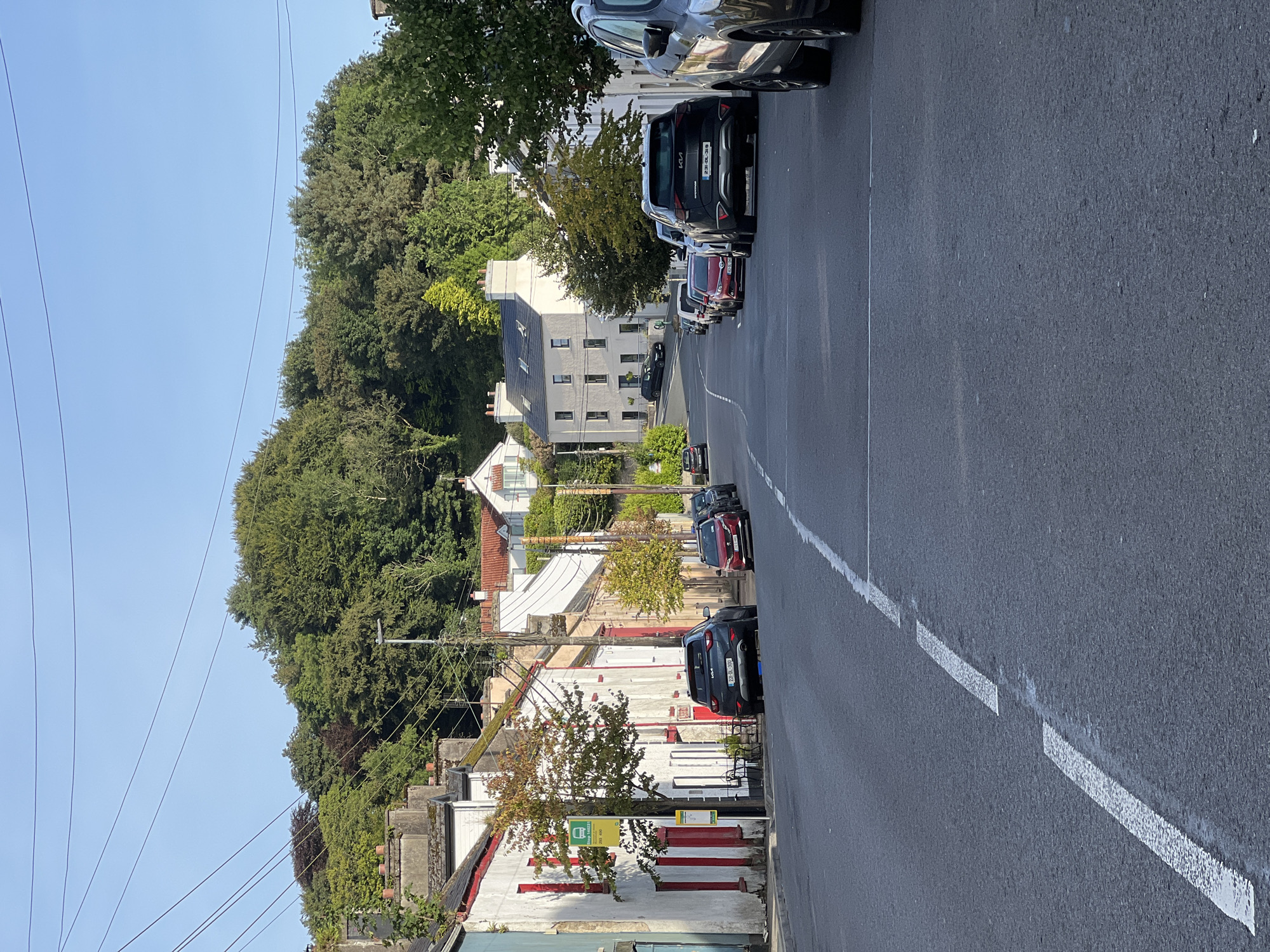 The image is a street-level, eye-level, vertical, full shot of a road in Mountcharles, Ireland, on a clear day with a blue sky. The road is paved and has white dashed lines marking the lanes. On the left side of the road, there are several terraced houses with white facades and red-painted window frames and doors. A yellow bus stop sign is visible. On the right side of the road, there are more houses, including a large cream-colored building with multiple windows and a tiled roof. Numerous parked cars are lined up along both sides of the street, mostly facing away from the camera. The background features a dense, green forested hill rising behind the houses. The overall impression is of a quiet, residential street in a town.
