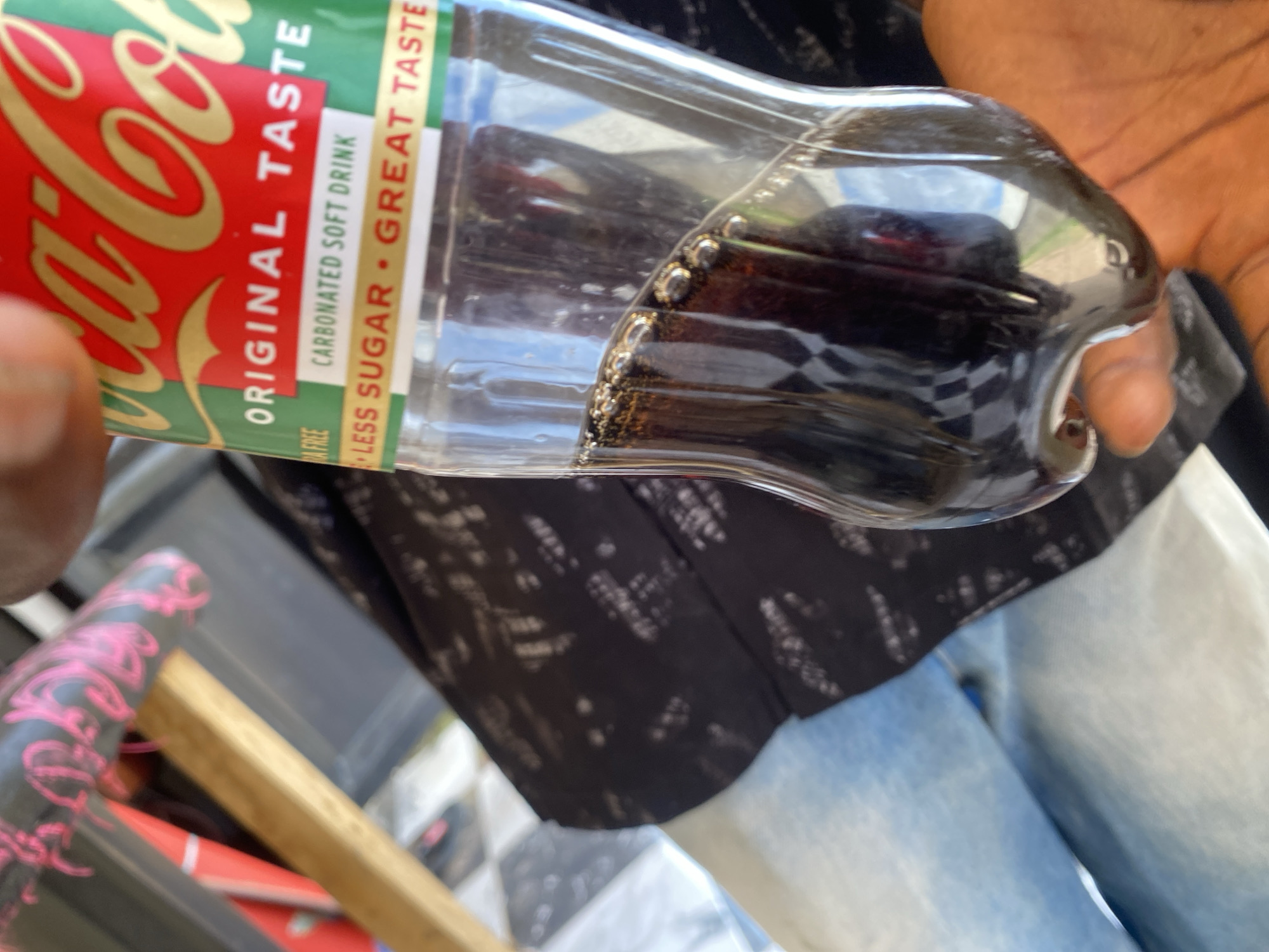 An extreme close-up shot captures a person's hand holding a partially filled bottle of Coca-Cola, with the drink's dark brown liquid and rising bubbles prominently displayed. The iconic Coca-Cola logo, featuring red, green, and gold lettering, is visible on the bottle's label. The background is blurred, but hints of denim fabric and what appears to be patterned clothing suggest an outdoor or casual setting. No specific landmarks or elements definitively place this scene in Jauro Yinu, Nigeria, as the image lacks contextual geographical markers.