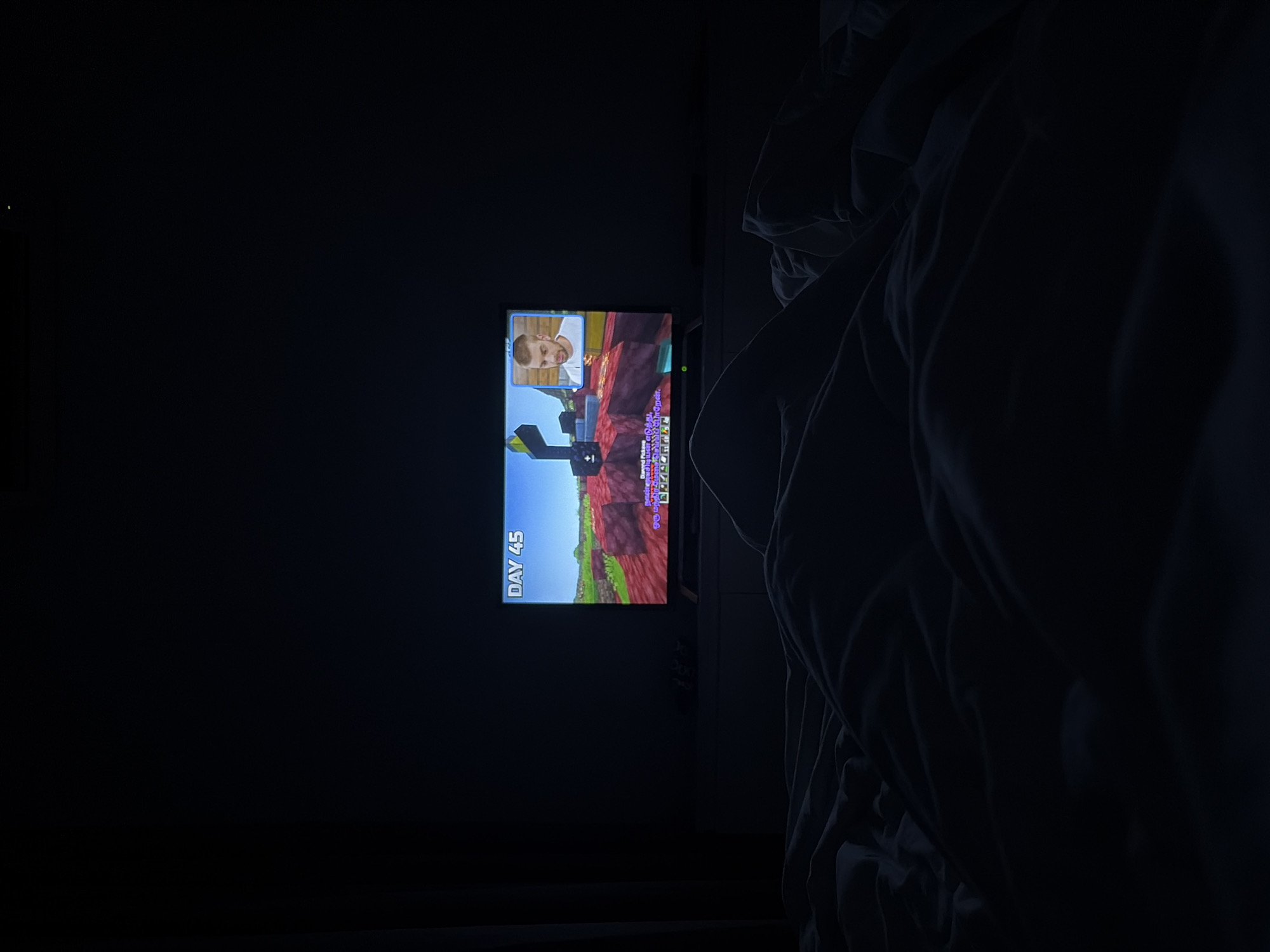 The image depicts a dark interior room, identified as being in Thủ Đức, Vietnam, viewed from a low perspective, likely from a bed. The dominant light source is a flat-screen television centered in the mid-ground, resting on a light-colored media console or dresser. The television screen displays a video game, characterized by blocky terrain with red and green elements and a distinctive structure of yellow and black blocks on the left. Prominently visible in the upper left corner of the screen is the text "DAY 45." In the lower portion of the screen, text includes "Diamond Pickaxe" followed by characters in the Tamil script, suggesting an in-game item or message, along with graphical icons representing inventory slots. A picture-in-picture window in the upper right corner of the television displays a close-up of a male individual with short, light hair and a beard, wearing a white t-shirt, appearing to react to the game content. In the foreground, dark-colored bedding, likely a duvet or blankets, is rumpled, indicating the presence of an unseen individual lying in bed, observing the television. The remainder of the room is obscured by darkness, with faint outlines of walls and potentially curtains on the left periphery.