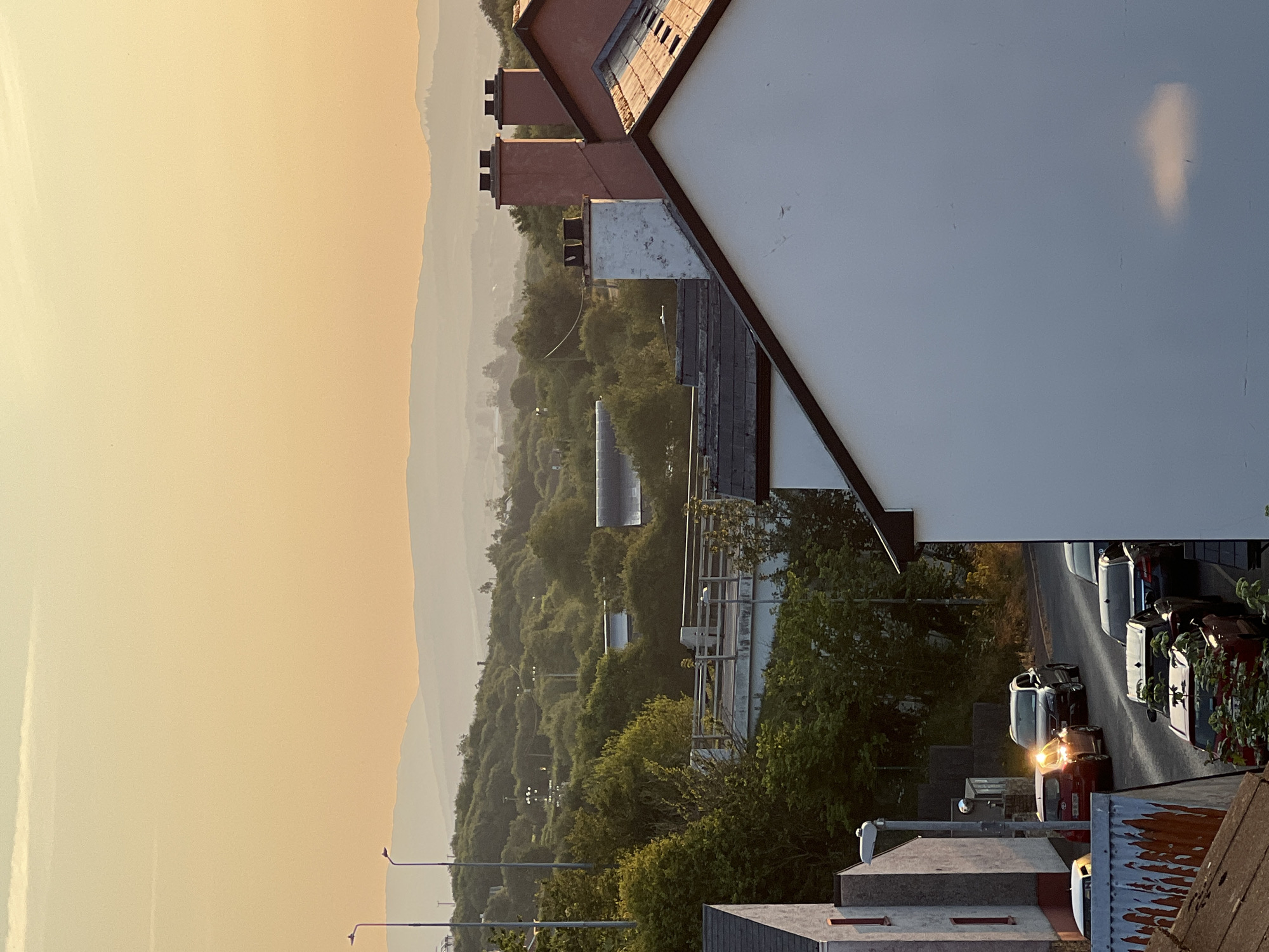 The image depicts a street scene in Mountcharles, Ireland, during the late afternoon or early evening, with the golden light of sunset casting a warm glow. In the foreground, the roofs and upper portions of houses are visible, one with a white facade and another with a reddish-pink exterior and multiple chimneys. A street stretches downhill, lined with parked cars and some in motion. A red hatchback is prominent, its headlights illuminated, and a dark SUV is behind it. Further down the street, additional vehicles are visible, including a black sedan.
The middle ground of the image is dominated by lush green trees and vegetation that covers rolling hills. A section of elevated infrastructure, possibly a bridge or walkway with railings, is visible above the trees. Streetlights are scattered throughout this area.
In the background, a series of hazy, layered hills recede into the distance, silhouetted against a soft, orange-tinted sky. A faint, elongated cloud is present in the upper portion of the sky. The overall impression is one of a quiet, suburban landscape bathed in the soft, diffused light of sunset.