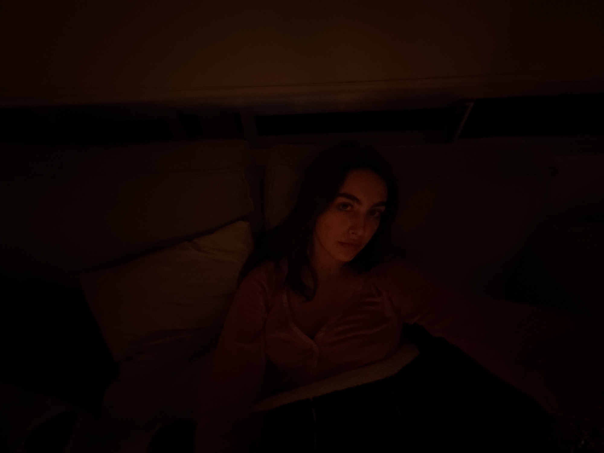 A person is lying on a bed in a dimly lit room. The person is facing forward and appears to be looking at the camera. The person is wearing a pink top and dark pants. A pillow is visible behind the person. The room appears to be indoors, but there is no identifiable information about the location in Mexico City. The lighting is poor, which makes it difficult to discern details. There are no visible actions or interactions.