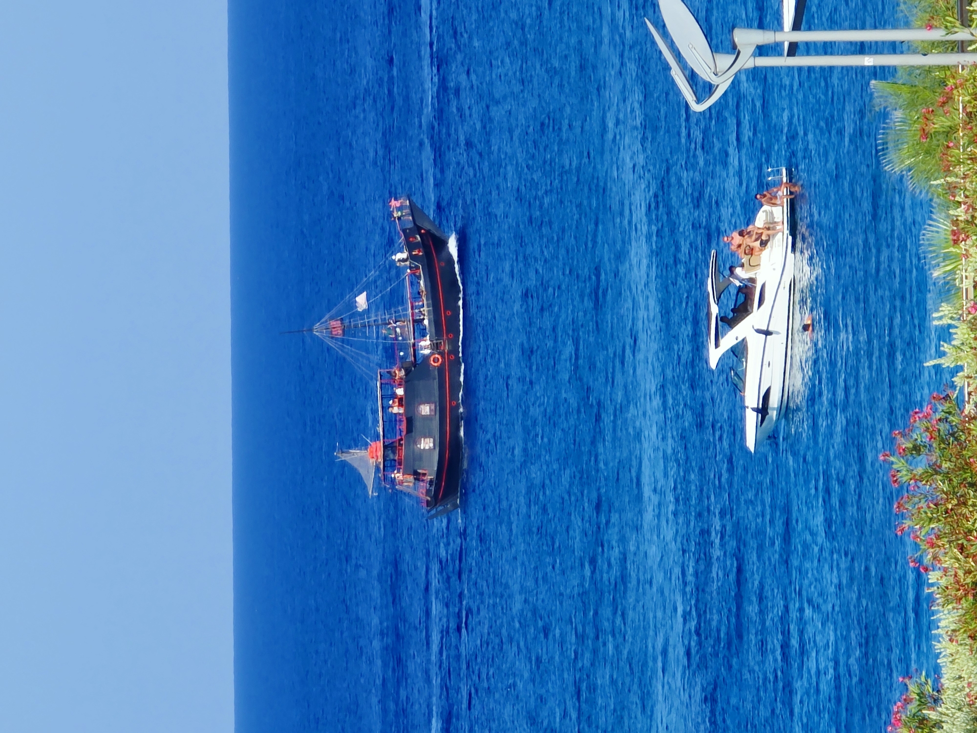 The image captures a bright, sunny day at sea, likely in the vicinity of Protaras, Cyprus, given the distinctive blue waters. In the upper half of the frame, a dark-colored pirate ship with red trim sails across the vibrant blue sea. The ship is populated with several individuals visible on its decks, some appearing to be enjoying the voyage. Below the pirate ship, a modern white speedboat glides through the water. This boat carries a group of people, some of whom are seated, while others are either in the water or preparing to enter it, suggesting a recreational activity. In the foreground, lush green foliage with vibrant pink flowers adds a touch of natural beauty to the coastal scene. A light-colored pole with a satellite dish is partially visible on the right side of the image, hinting at nearby infrastructure or residential buildings. The clear blue sky above completes the serene coastal panorama.