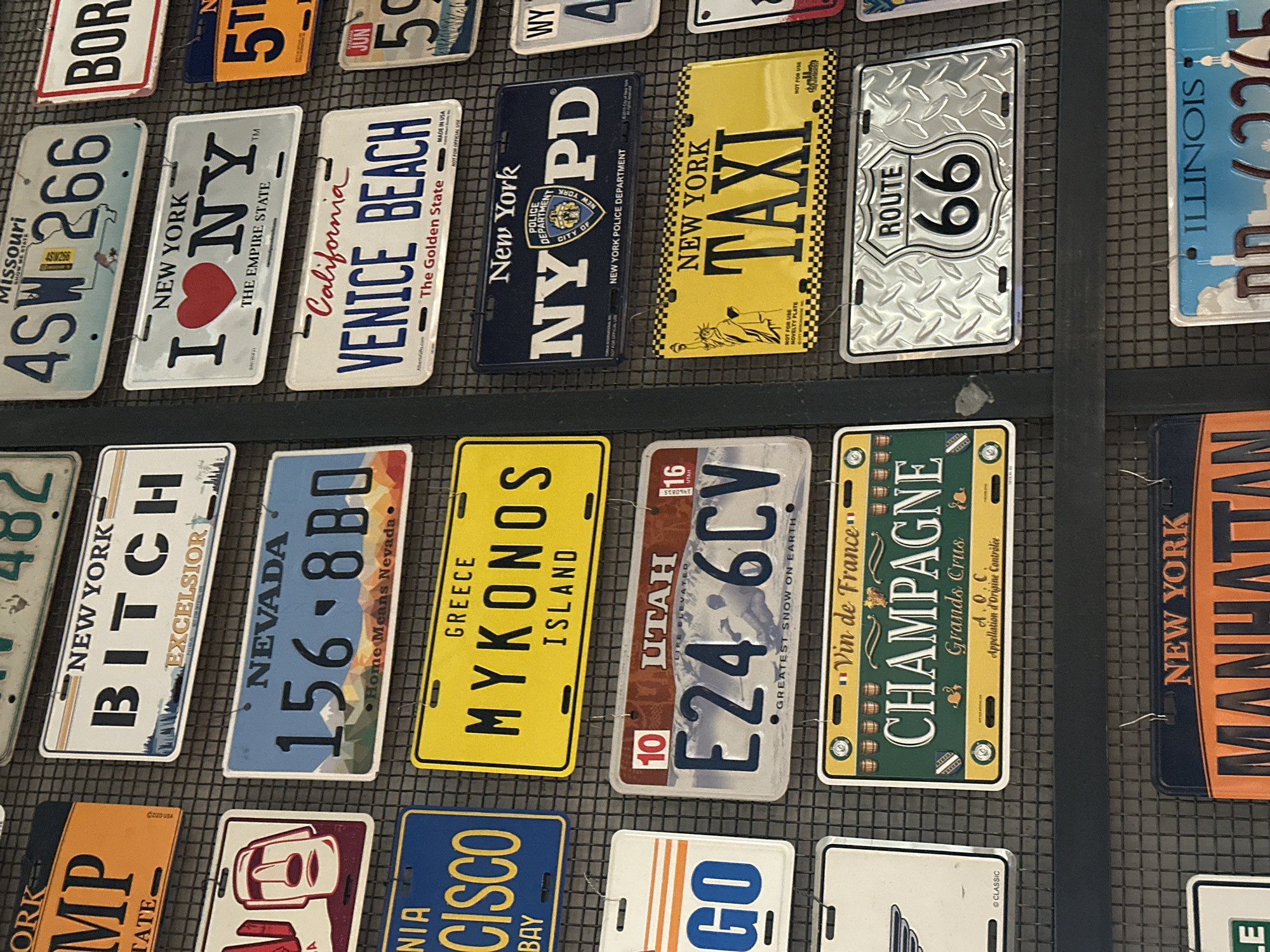 The image shows a collection of novelty license plates displayed on a wire grid. The plates represent various locations and themes, including US states like Missouri, New York, California, Nevada, Utah, and Illinois. There are also plates referencing cities such as Venice Beach, Cisco Bay, and Manhattan, as well as themes like Route 66, taxi, and champagne. There is no information regarding the location in the city of Krasnoyarsk, Russia.