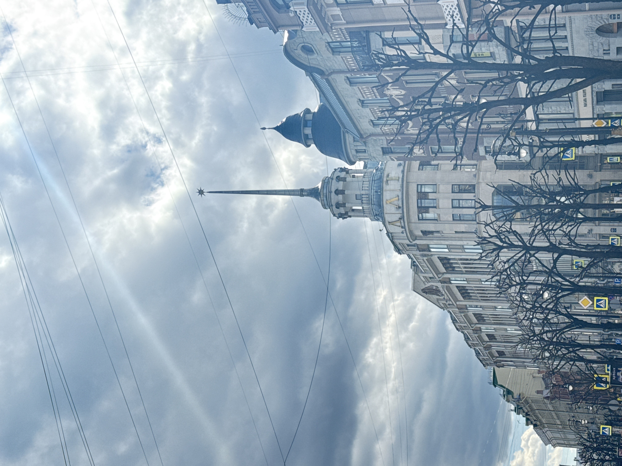 The image captures a street scene in Saint Petersburg, Russia, with a distinctive architectural skyline dominated by a tall spire topped with a star. The building's facade is visible, featuring multiple windows and intricate detailing. A smaller, rounded turret with a dark-colored roof is also prominent. Bare trees with intricate branches frame the buildings, adding a stark contrast against the overcast sky. Power lines stretch across the sky, intersecting with the spire. Street signs are visible along the side of the street, contributing to the urban environment. The sky is filled with gray clouds, suggesting a somewhat overcast or potentially stormy weather.