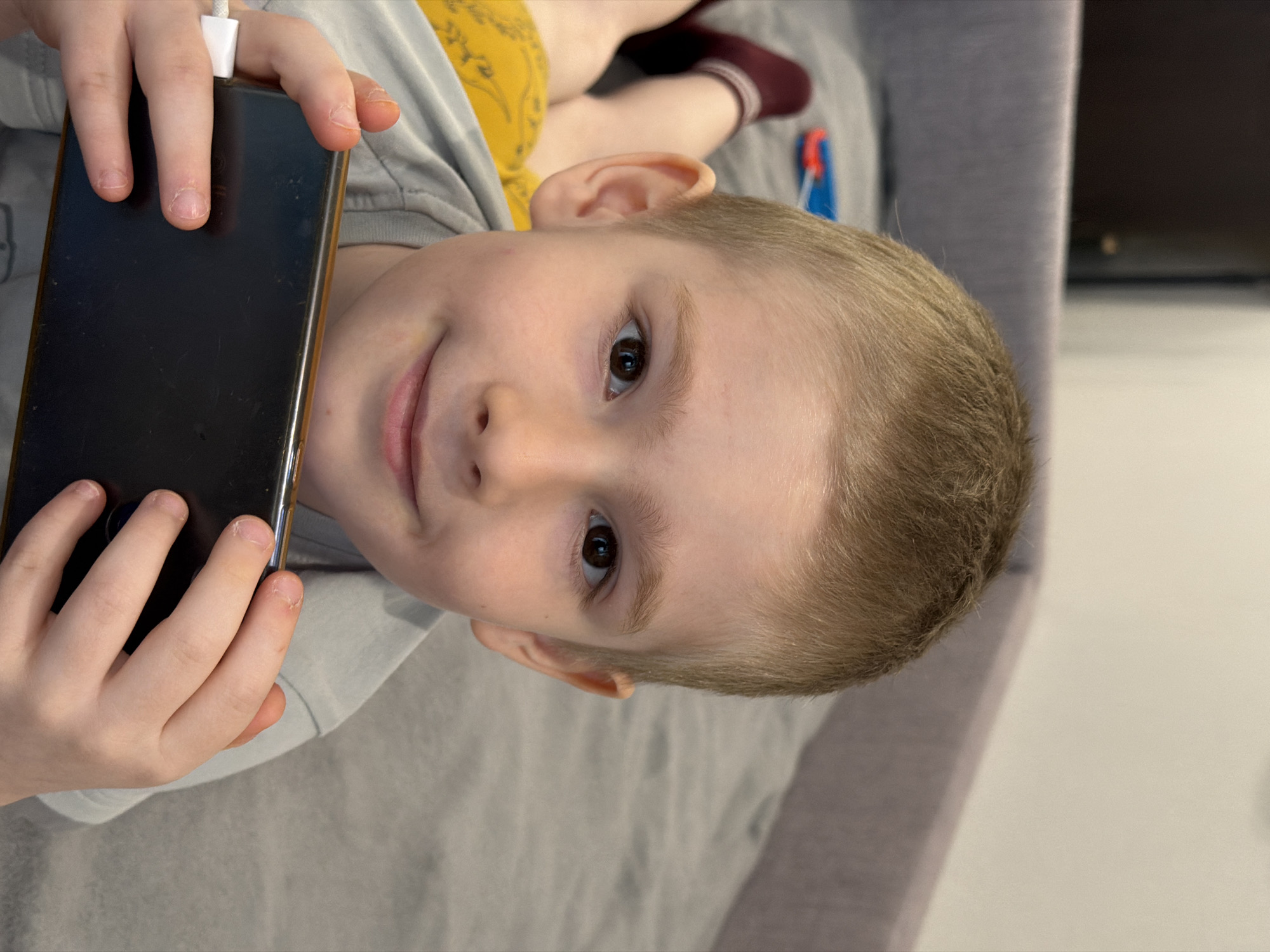 The image displays a close-up portrait of a young child, likely a boy, with fair skin, light brown hair, and dark brown eyes. He is lying down on a grey upholstered surface, looking directly at the camera with a soft, engaging smile. His head is positioned towards the left side of the frame.
The child holds a dark-colored smartphone vertically with both hands in front of him. A white charging cable is plugged into the top of the phone, indicating it is being charged. His fingers are visible, with natural, closely trimmed fingernails.
In the upper right portion of the background, parts of another person's body are visible, including a leg wearing a yellow garment (possibly shorts) and a foot adorned with a dark red sock, suggesting another individual is also relaxing nearby. The setting appears to be an indoor home environment, possibly a living room or bedroom, indicated by the upholstered furniture and subtle hints of room elements in the background. The overall impression is one of relaxed contentment. There is no visible text present in the image. The location context for this scene is Засечное, Russia.