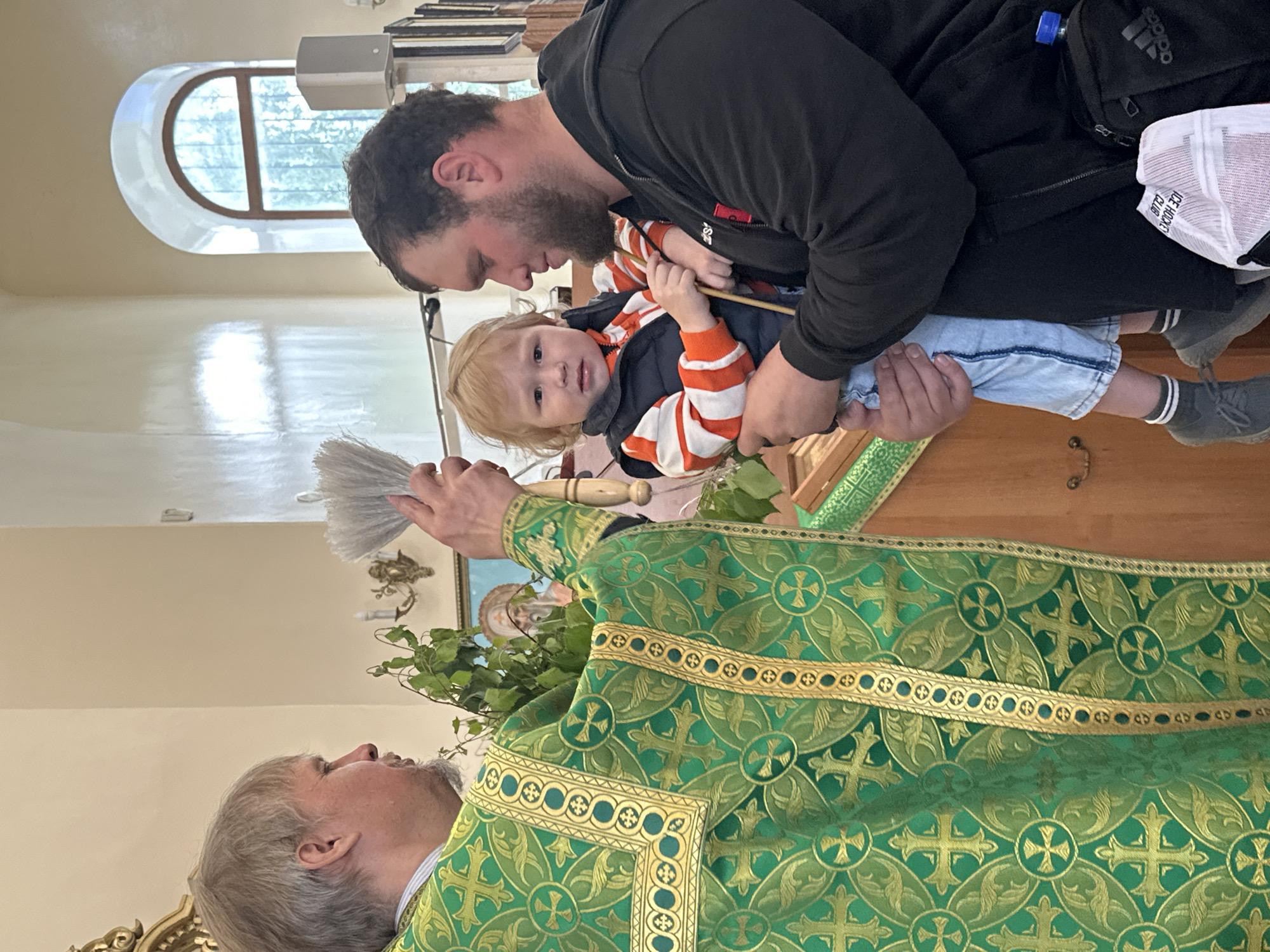 The image shows an Eastern Orthodox priest in Votkinsk, Russia, performing a blessing or ritual. The priest, wearing a green and gold robe, holds a bundle of leaves in his left hand and a grey brush-like object in his right. Facing him is a man holding a young child, likely a toddler, in his arms. The man is wearing a black jacket, an orange and white striped shirt, and light blue jeans. The child is looking directly at the camera. In the background, there is a rounded window, a piece of religious art, and other church-related items. The interior appears to be a church or religious building in Votkinsk.