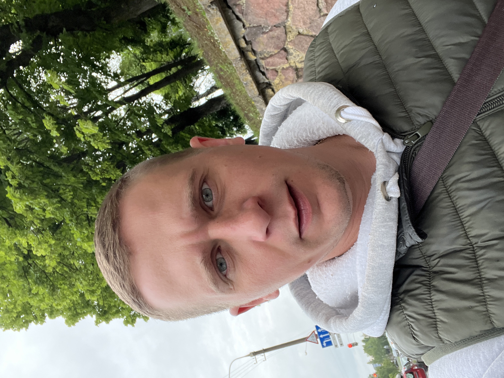 A male individual with light brown hair and blue eyes is captured in a close-up selfie. He is wearing a white hoodie layered with a green puffer vest. The background features lush green trees and a cloudy sky, suggesting an outdoor urban setting. A stone wall and a street sign are visible, and traffic lights are dimly lit in the distance, hinting at a street-side location. The man's expression is neutral. The image appears to be taken in the city of Minsk, Belarus, given the context of the street signs and general environment.