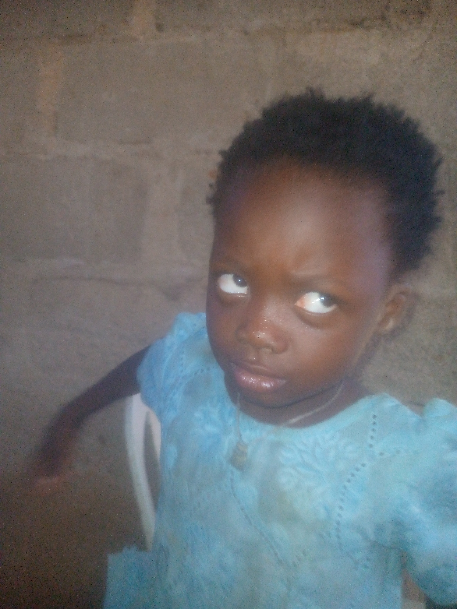 A young, dark-skinned child with short, dark hair and a light blue lacy shirt is the primary subject. She is looking upwards and to the left with a somewhat serious expression. The background is a rough, grayish wall. The location appears to be indoors.