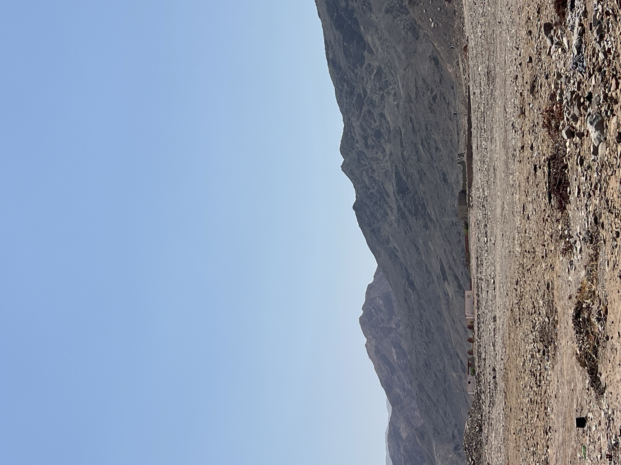 In the city of Unknown, Egypt, a photograph captures a desolate, rocky landscape under a clear blue sky. Jagged, dark mountains dominate the background, their slopes etched with the texture of time and erosion. In the mid-ground, a collection of low-lying, light-colored buildings with red-tiled roofs are visible, appearing to be situated within the rough terrain. The foreground is a chaotic expanse of stones, gravel, and debris, suggesting an undeveloped or abandoned area. The overall impression is one of a harsh, arid environment, with human presence indicated only by the distant structures.