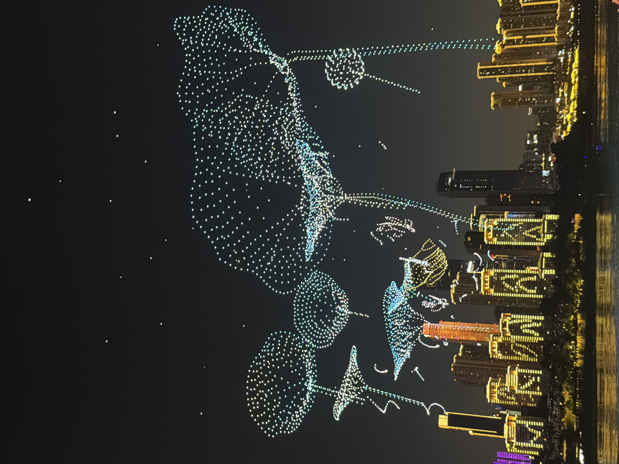 An aerial drone light show is taking place over the Nan'an District in China. Numerous drones, forming intricate shapes of various objects, are suspended in the dark night sky. These objects include what appear to be large, luminous dandelion-like structures and possibly other floral or abstract forms, all composed of countless bright, white and blue dots of light. Below the drone display, a cityscape is visible, illuminated by the lights of numerous tall buildings. The buildings are a mix of modern skyscrapers and apartment complexes, many of which are outlined and decorated with bright yellow and some purple lights, creating a vibrant urban landscape. The reflection of these lights can be seen on the surface of a body of water in the foreground. The overall scene depicts a festive and technologically advanced display over an urban setting.