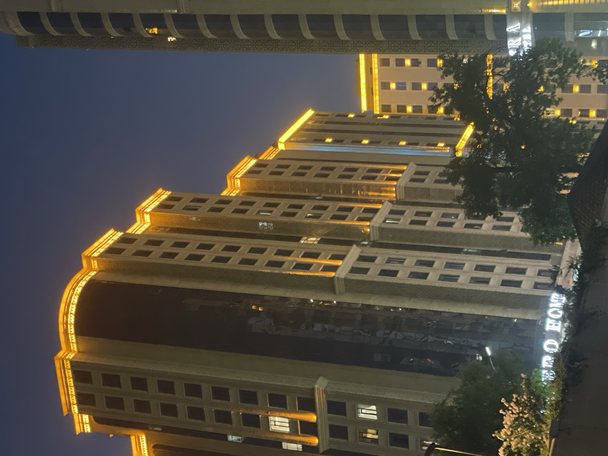 This photograph captures a view of modern high-rise buildings in Dushanbe, Tajikistan, at dusk or night. The buildings are illuminated with prominent orange-yellow lighting along their upper facades, highlighting architectural details and creating a warm glow against the deep blue sky. The architecture features a repetitive pattern of windows and decorative elements. In the foreground, a street light and some foliage, including a bush with pink flowers, are visible. A sign with white illuminated lettering that reads "EXPO HOME" is present on a lower structure. To the right, another building is partially visible with similar architectural styling and lighting. The overall impression is one of urban development and nighttime illumination in a city.