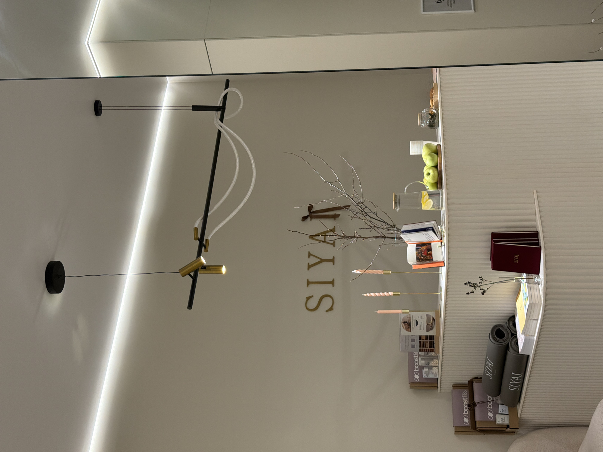 The image displays the reception or retail area of the SIYAI yoga and stretching studio, located at Bolshaya Bronnaya Street, 23с1, Moscow, Russia. The scene features a clean, modern interior with light-colored walls and a ceiling illuminated by linear LED strip lighting. A contemporary ceiling light fixture with black rods, golden cylindrical spotlights, and white looped tubing hangs centrally. On the wall above a light-colored, ribbed counter, golden three-dimensional letters spell "SIYAI." The counter is arranged with a dry tree branch centerpiece adorned with a brown ribbon, two pink spiral candles in gold holders, and several open books. To the left, rolled grey yoga mats bearing the "SIYAI" logo are displayed next to "boostlife" branded boxes and bottles. A lower shelf on the counter holds additional stacked books or brochures. To the right of the centerpiece, a glass pitcher with lemon-infused water, a pile of green apples, white paper cups, a glass jar containing dried botanicals or tea leaves, and a wooden plate with small snacks are arranged. A large glass panel or mirror is visible on the far right, reflecting the overhead lighting. No individuals are present in the image.