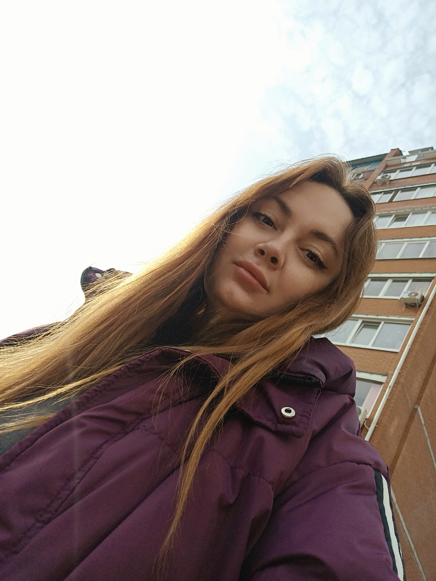 A young female, appearing in her late teens or early twenties, is depicted from a low-angle perspective, looking directly into the camera. She has long, light brown hair falling over her shoulders and is wearing a deep purple, possibly padded, jacket. The jacket features visible stitching, a silver snap button on a front flap on her right side, and a white and black stripe detail on the upper right arm. The background reveals a bright, overcast sky to the upper left. To the right, a multi-story residential building with reddish-brown brick walls is visible, featuring multiple white-framed windows and at least one external white air conditioning unit mounted below a window. The top right corner shows a portion of the building's dark-colored, sloped roof. The scene is consistent with an urban environment in the city of Chaikovsky, Russia, though a precise location within the city cannot be identified from this image.