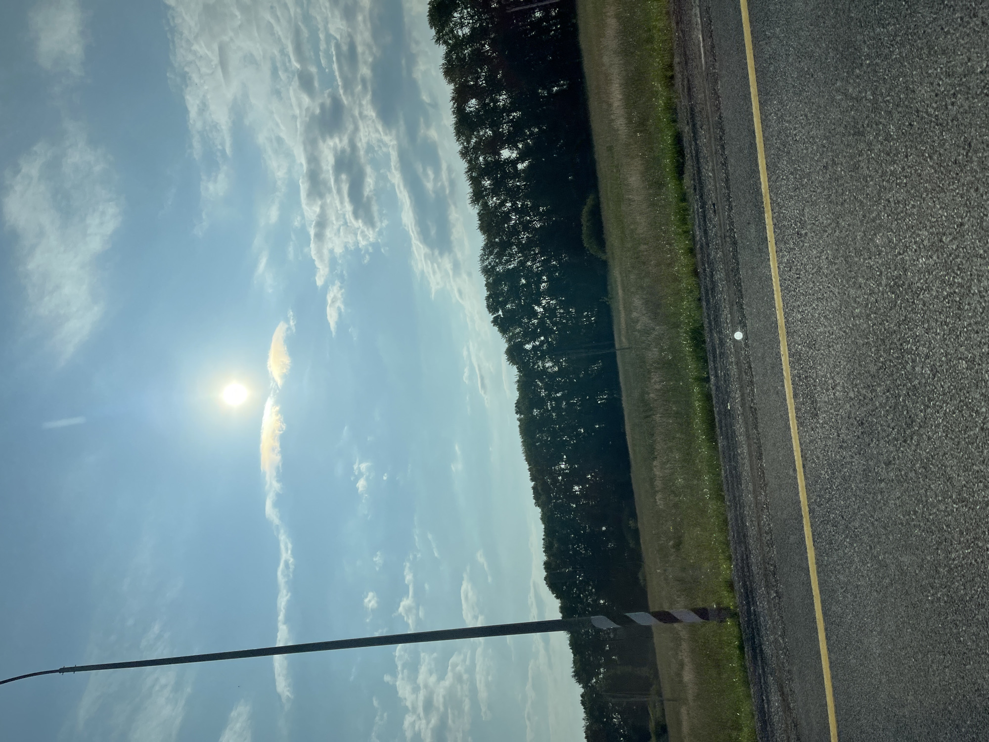 The image depicts a roadside view under a bright, partly cloudy sky. In the foreground, a portion of an asphalt road with a yellow dividing line is visible. To the left, a tall pole with red and white diagonal markings stands near the road. Beyond the road, a field of dry grass stretches towards a dense line of trees forming a dark, silhouetted background. The sun is high in the sky, creating a bright glare with a halo effect around it, and casting light on some of the clouds. The scene appears to be a rural or semi-rural area, possibly on the outskirts of a city or town. No people or vehicles are present in the image. The overall impression is one of a sunny day in a natural landscape setting.