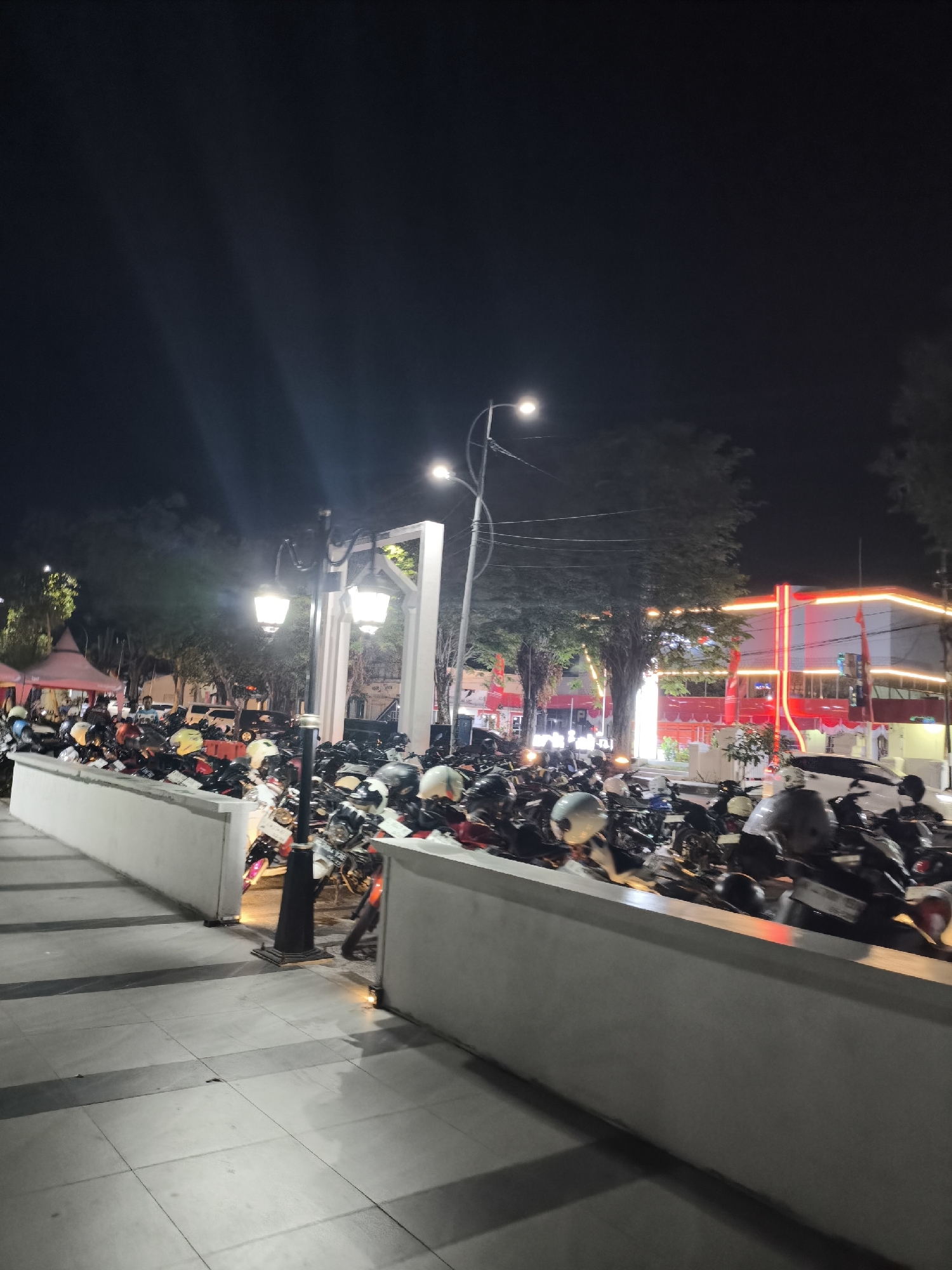 This is a low-angle, night-time photograph taken in Lumajang, Indonesia, likely at a public gathering or event. The scene is dominated by a large number of parked motorcycles filling a designated area. The motorcycles are mostly covered with helmets, indicating recent activity. In the background, a brightly lit building with red and white accents, possibly a commercial establishment or government office, is visible. Several trees with illuminated foliage are scattered throughout the scene, and there are streetlights providing illumination. A white concrete barrier runs along the foreground, separating a pedestrian walkway from the motorcycle parking area. The overall impression is that of a busy night in a public space, with a focus on the parked vehicles.