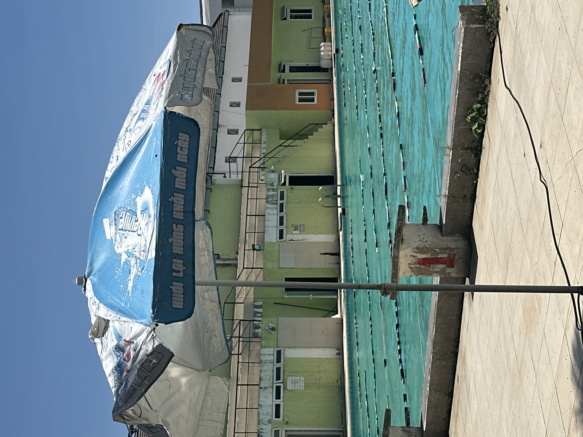 A partially visible swimming pool is adjacent to a three-story building, while an open advertising umbrella is in the foreground. The building is painted in light shades of green and brown, with white windows and black staircases. The pool water is a vibrant light blue color. The advertisement on the umbrella is a combination of white and blue, with prominent lettering in Vietnamese. The scene is in Nha Trang Ward, Vietnam.