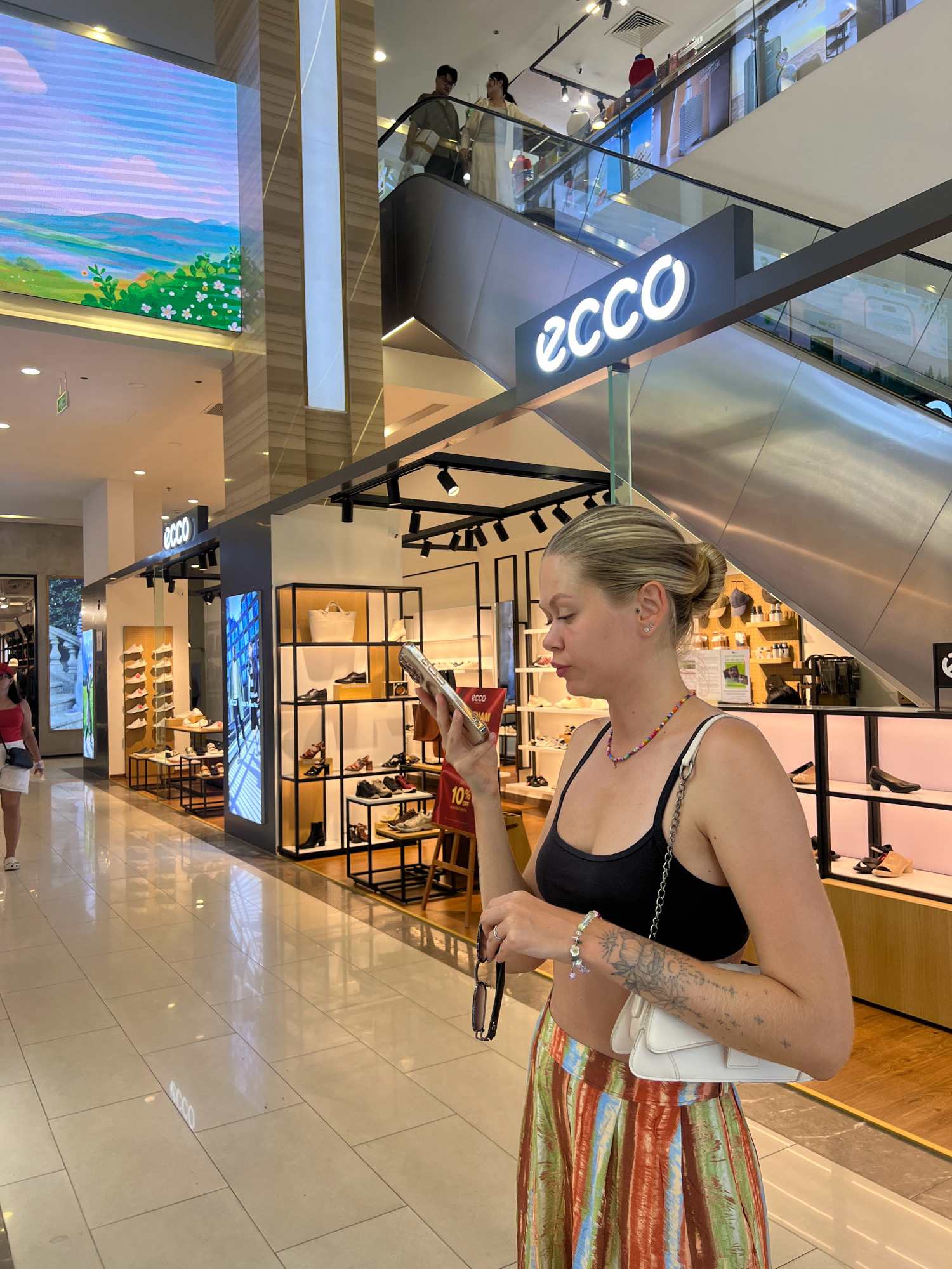 In Nha Trang, Vietnam, a blonde woman with her hair in a bun is standing in a brightly lit shopping mall, looking at her phone. She is wearing a black crop top, a colorful striped skirt, and a beaded necklace. A white purse hangs from her shoulder, and she holds a pair of sunglasses in her hand. The mall features an escalator in the background with a prominent "ECCO" sign. Various shops are visible, including a shoe store displaying rows of footwear on shelves. The floor is tiled and reflects the ambient light.