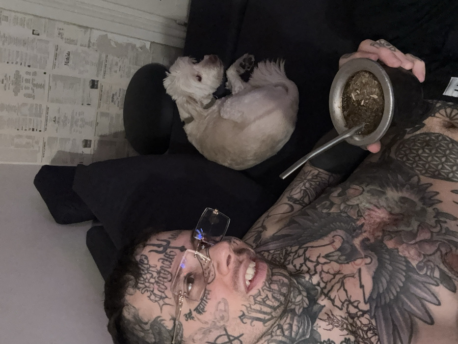 An indoor scene depicts an adult male and a small dog on a dark-colored couch. The male, positioned on the left side of the frame, is heavily tattooed across his face, neck, chest, and arms, with prominent script and symbolic designs visible. He wears rimless glasses with gold-colored accents and looks directly towards the viewer with a slightly open mouth. In his left hand, he holds a dark mate gourd with a silver-colored rim, containing yerba mate and a metal bombilla. To the male's right, a light-colored, short-haired dog, possibly a terrier mix, is curled up on the couch, wearing a dark collar, and facing away from the male's direct line of sight. The background wall is light-colored and partially covered with multiple sheets of aged paper, resembling newspaper clippings or documents. The specific location within the city of Buenos Aires, Argentina, cannot be determined from the visual information provided.