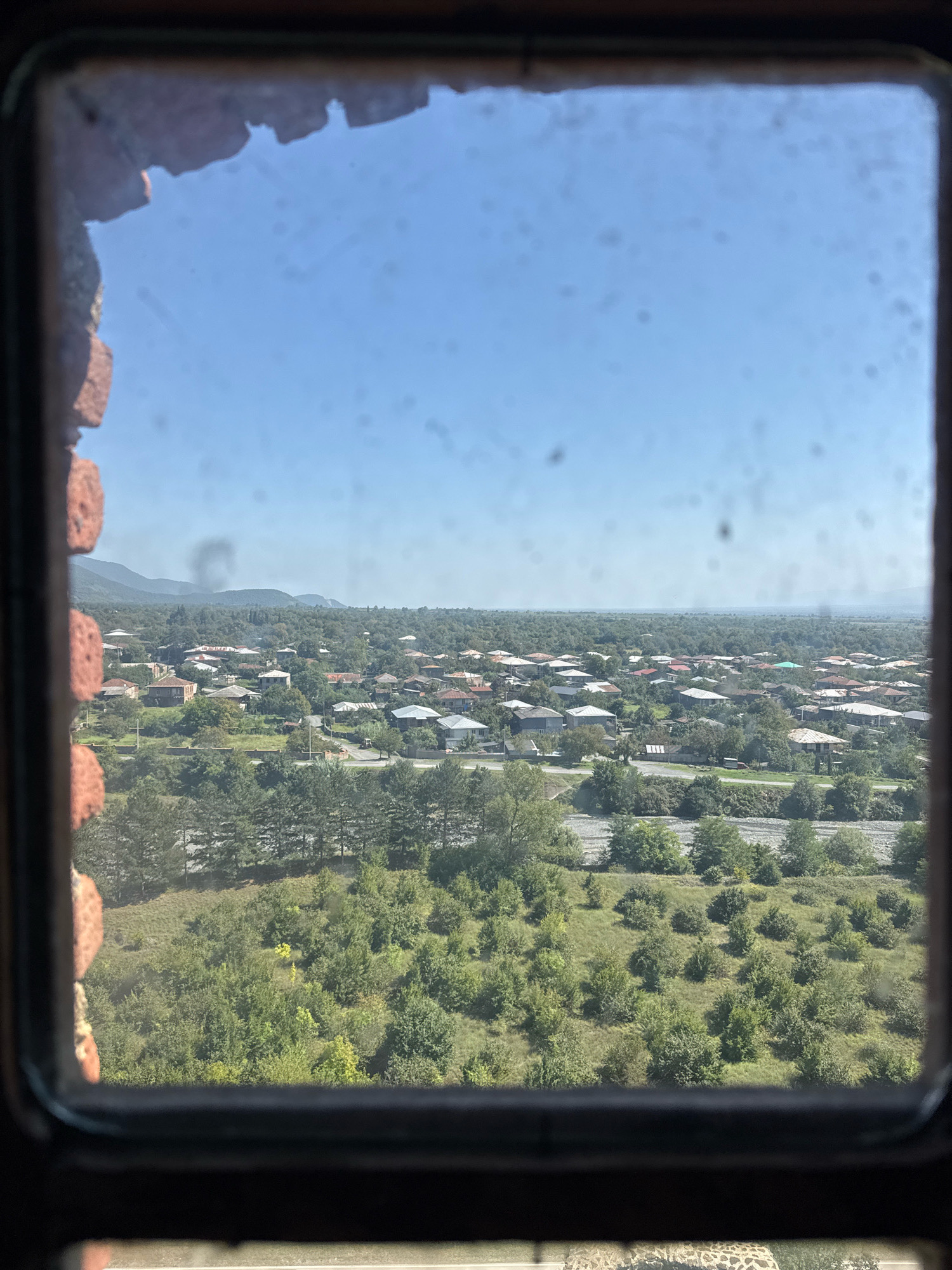 The image, taken from inside a rough-hewn brick structure through a dusty window, presents a view of the city of Eniseli, Georgia. The foreground is dominated by the interior of the structure, with visible, weathered bricks forming the frame of the window. Beyond the window, a panorama unfolds. A dense expanse of green trees and vegetation occupies the middle ground, interspersed with a settlement of houses. These houses are predominantly single-story with red-tiled roofs, clustered together, suggesting a residential area. A road or path is visible winding through the greenery, with a few cars present. In the background, rolling hills or mountains form a natural backdrop, partially obscured by the hazy atmosphere. The sky is a clear, bright blue, with no clouds visible. The overall impression is a rural or semi-rural landscape bathed in daylight.