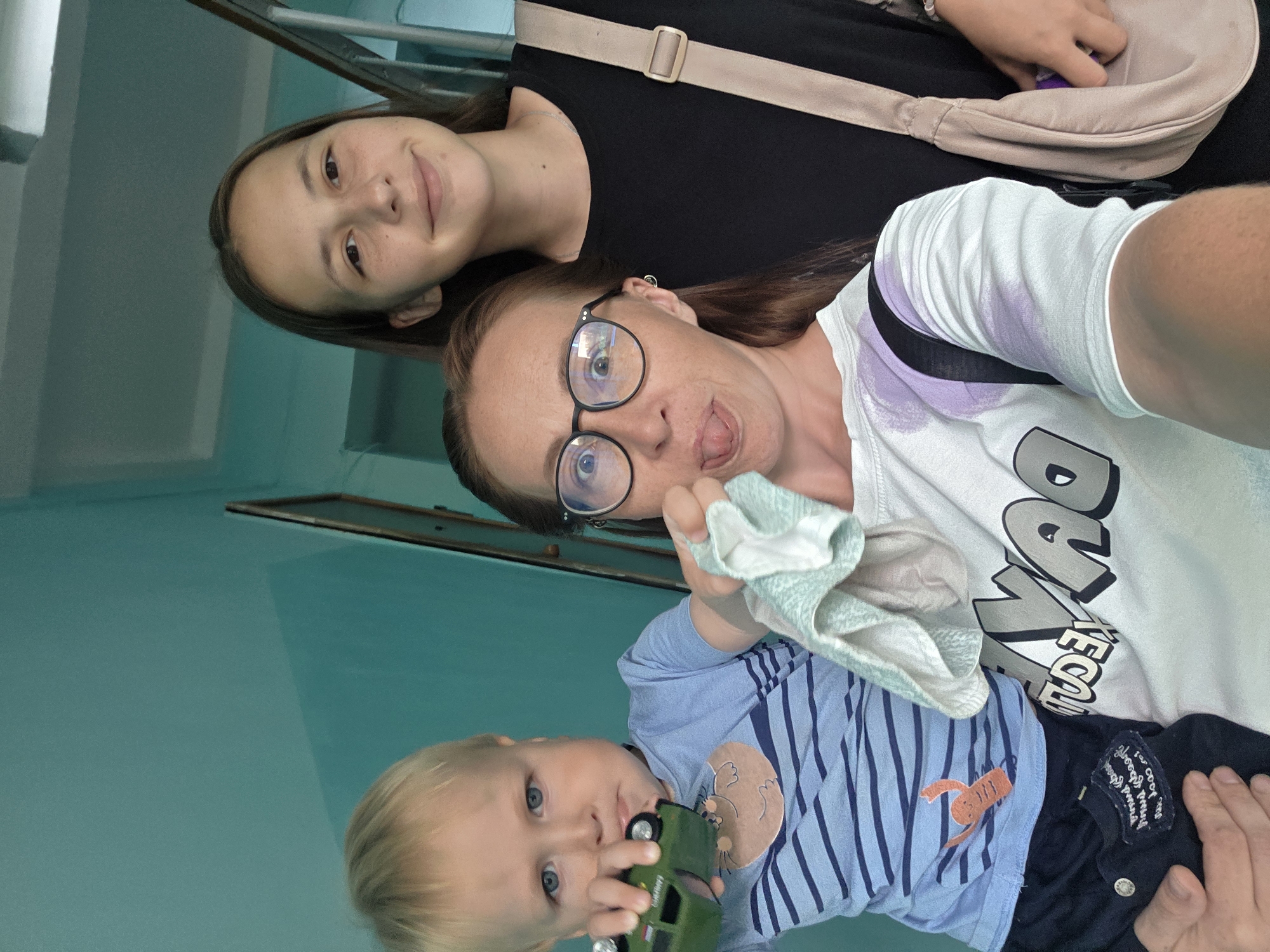 Three individuals, a young boy and two females, are posing for a selfie indoors. The background suggests a residential building hallway, possibly in Chaikovsky, Russia, with teal-colored walls and a staircase visible on the right.
The young boy, in the foreground and to the left, has blond hair and blue eyes. He is holding a green toy car and wearing a blue striped t-shirt with an orange lobster graphic. He is being held by the female in the center.
The central female has reddish-brown hair and is wearing glasses and a white t-shirt with a graphic design. She is sticking her tongue out and holding a light blue and white cloth.
The female on the right has brown hair and is wearing a black t-shirt. She has a light beige crossbody bag slung over her shoulder. Her right hand is visible reaching into the bag.
The lighting is moderate, and the overall scene captures a candid family moment.