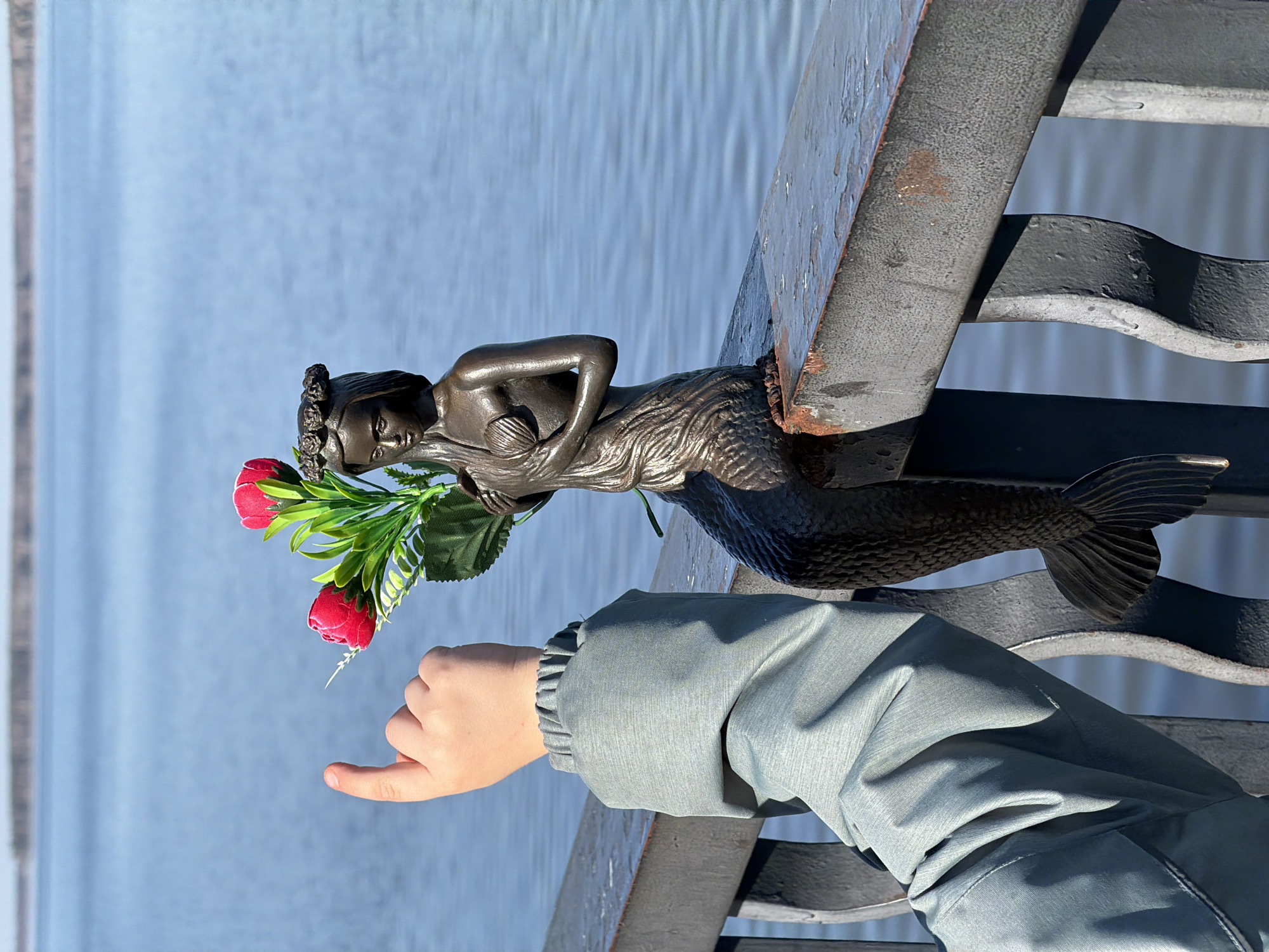 A metal sculpture of a mermaid holding red flowers is seen along a metal railing. A person's hand in a jacket is reaching towards the flowers. The backdrop is a body of water. The scene is located in the city of Chaikovsky, Russia.