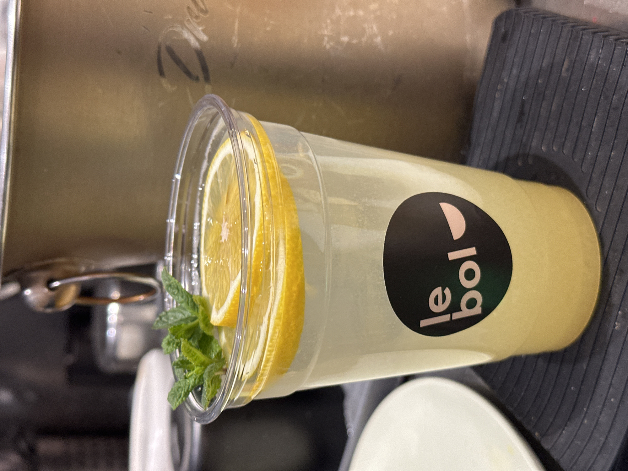A clear plastic cup of lemonade with lemon slices and mint leaves sits on a textured black surface. The cup has a logo on the side, with "le bol" written vertically. The background shows parts of a metal appliance and a white plate. The location is in Novi Sad, Serbia.