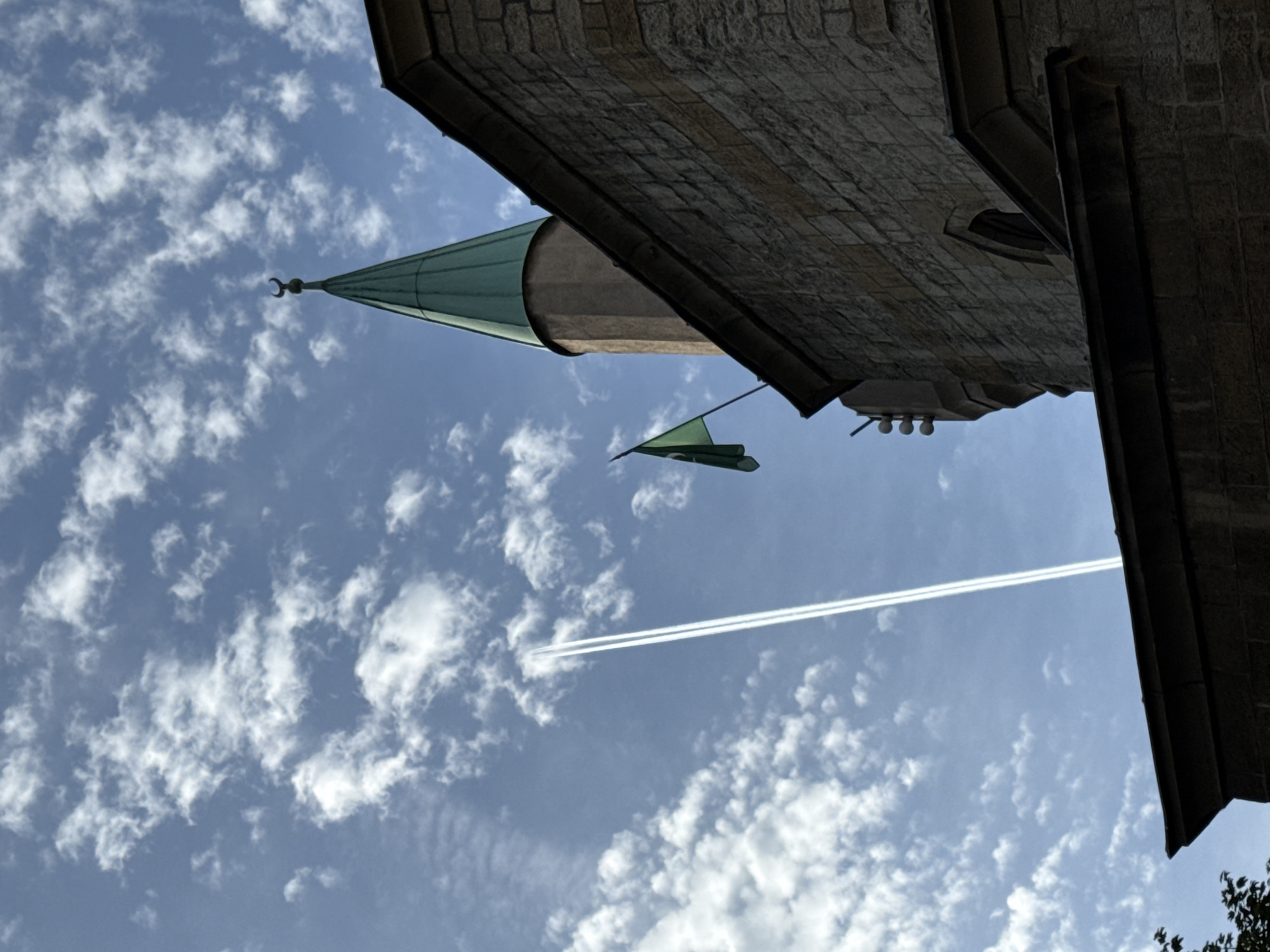The image is a low-angle shot looking up at the sky, partly obscured by the corner of a building. The building appears to be constructed of brick or stone. To the left, a green, conical roof with a crescent moon finial is visible, indicating it might be part of a mosque. A green flag hangs from the building. The sky is blue with scattered cumulus clouds. A jet contrail stretches across the sky. The location is likely the Bajrakli Mosque in Belgrade, Serbia.