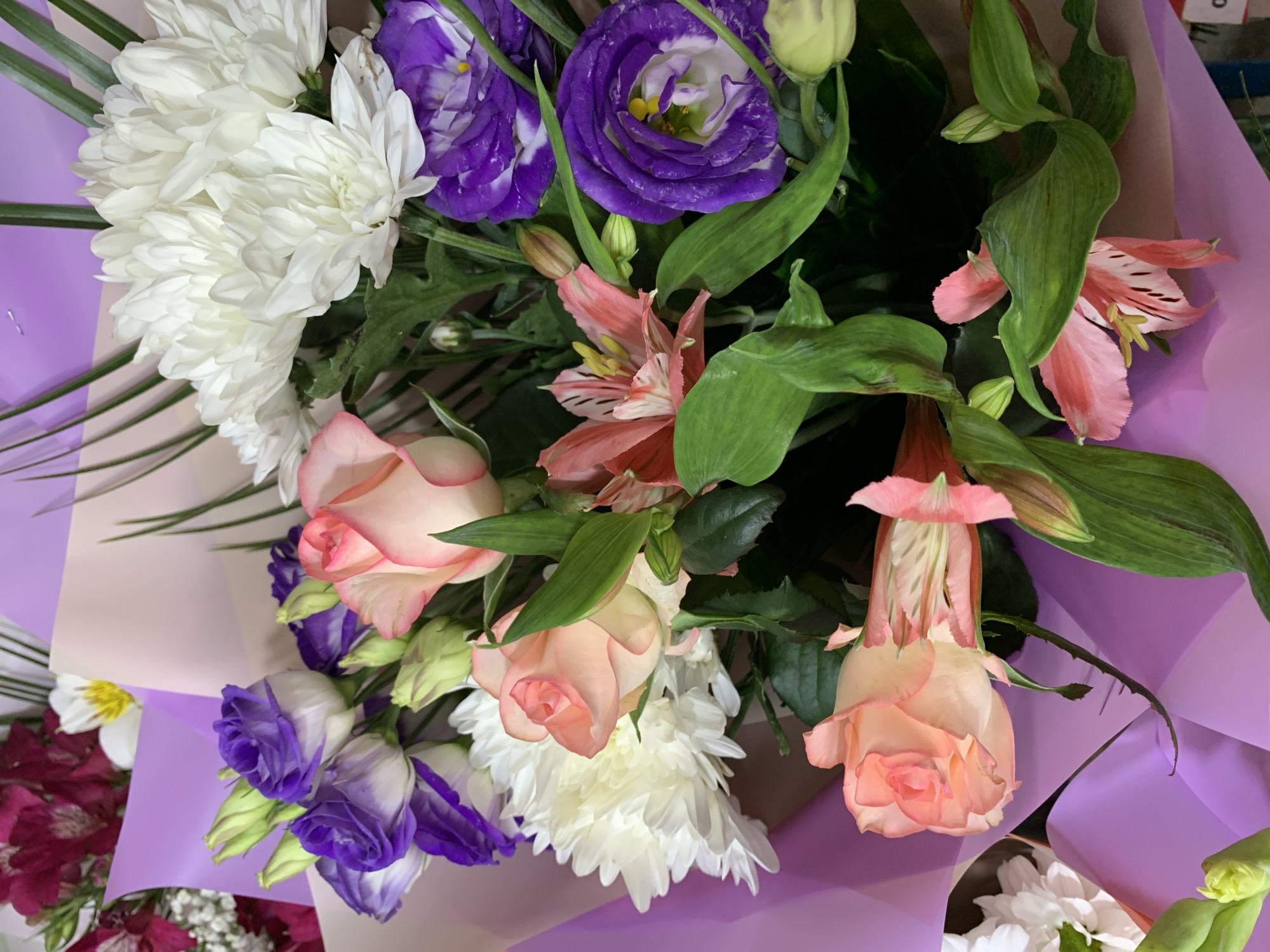 A close-up shot captures a vibrant bouquet of flowers. The arrangement features a variety of blooms including peach roses, white chrysanthemums, purple lisianthus, and pink alstroemeria, all nestled amongst lush green foliage. The flowers are wrapped in pastel purple and pink paper. The scene is indoors, with no identifiable external environmental features present that would indicate a specific location within the city of Sumy, Ukraine, or any other geographical area. The lighting suggests artificial indoor illumination. No people or specific actions are depicted.