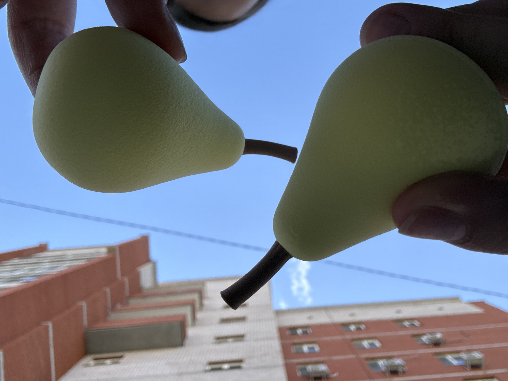 Two light green, artificial pears with short, dark brown stems are held by human fingers against a bright, clear blue sky. The top pear is grasped by a thumb and forefinger on its left side, while the bottom pear is held similarly by different fingers on its right side. A small, out-of-focus portion of a person's face, likely the chin or jawline, is visible on the far right edge of the image.
In the background, positioned diagonally from the lower-left to the upper-left, is a multi-story building located in the city of Chaikovsky, Russia. The building features reddish-brown colored balconies or architectural trim and lighter-colored (possibly white or beige) wall sections with numerous rectangular windows. The building appears slightly out of focus relative to the pears. A thin, dark line, possibly a wire or cable, spans diagonally across the sky from the upper-left to the lower-right, passing behind the top pear. The overall composition suggests an upward-facing perspective with strong backlighting from the sky.