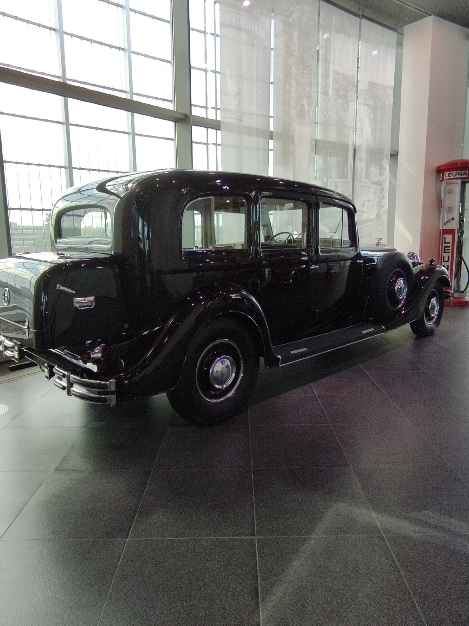 The image shows the rear three-quarter view of a vintage black sedan parked on a tiled floor in a museum. The car has a spare tire mounted on the rear passenger side fender. To the right of the car is a red and white vintage gasoline pump with the brand "LEUNA" visible. The background features a large window with a grid pattern and sheer white curtains. The location is identified as the Audi Forum Ingolstadt in Ingolstadt, Germany. There are no people visible in the image.