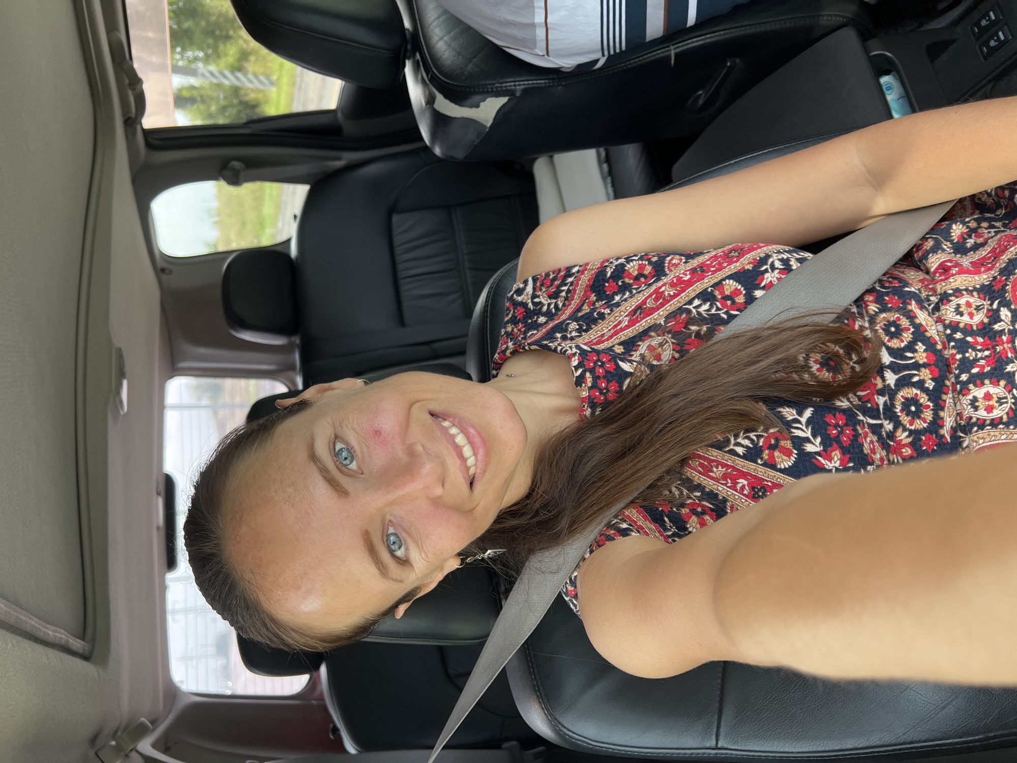 A woman with blue eyes, brown hair, and fair skin smiles at the camera from the driver's seat of a car. She is wearing a red, blue, and gold patterned sleeveless dress and a gray seatbelt. Her right arm is extended towards the camera, holding it. The interior of the car is visible, with black leather seats and a sunroof. The background is blurred, showing greenery and what appear to be power lines. The woman's left earlobe has a silver earring shaped like a dagger. She has a small mole or blemish on her right cheek. This image was taken in Anfalovo, Russia.