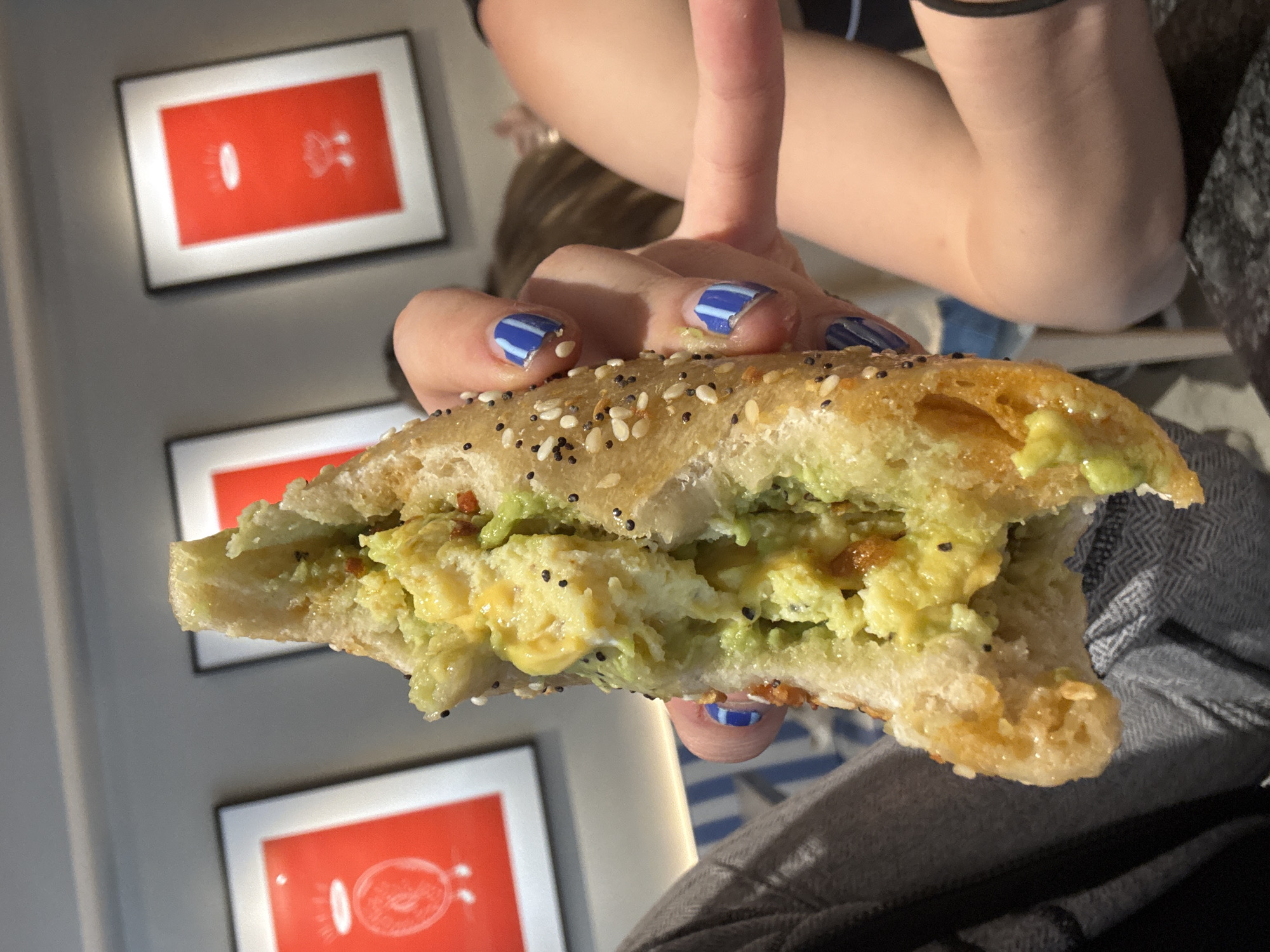 A person is holding a bagel sandwich with avocado and eggs in front of a wall with three framed red artworks. The bagel is topped with sesame and poppy seeds. The person has blue and white striped nail polish. The location is likely in Mexico City, Mexico, based on the presence of the artwork, which is a common theme in cafes and restaurants in the city.