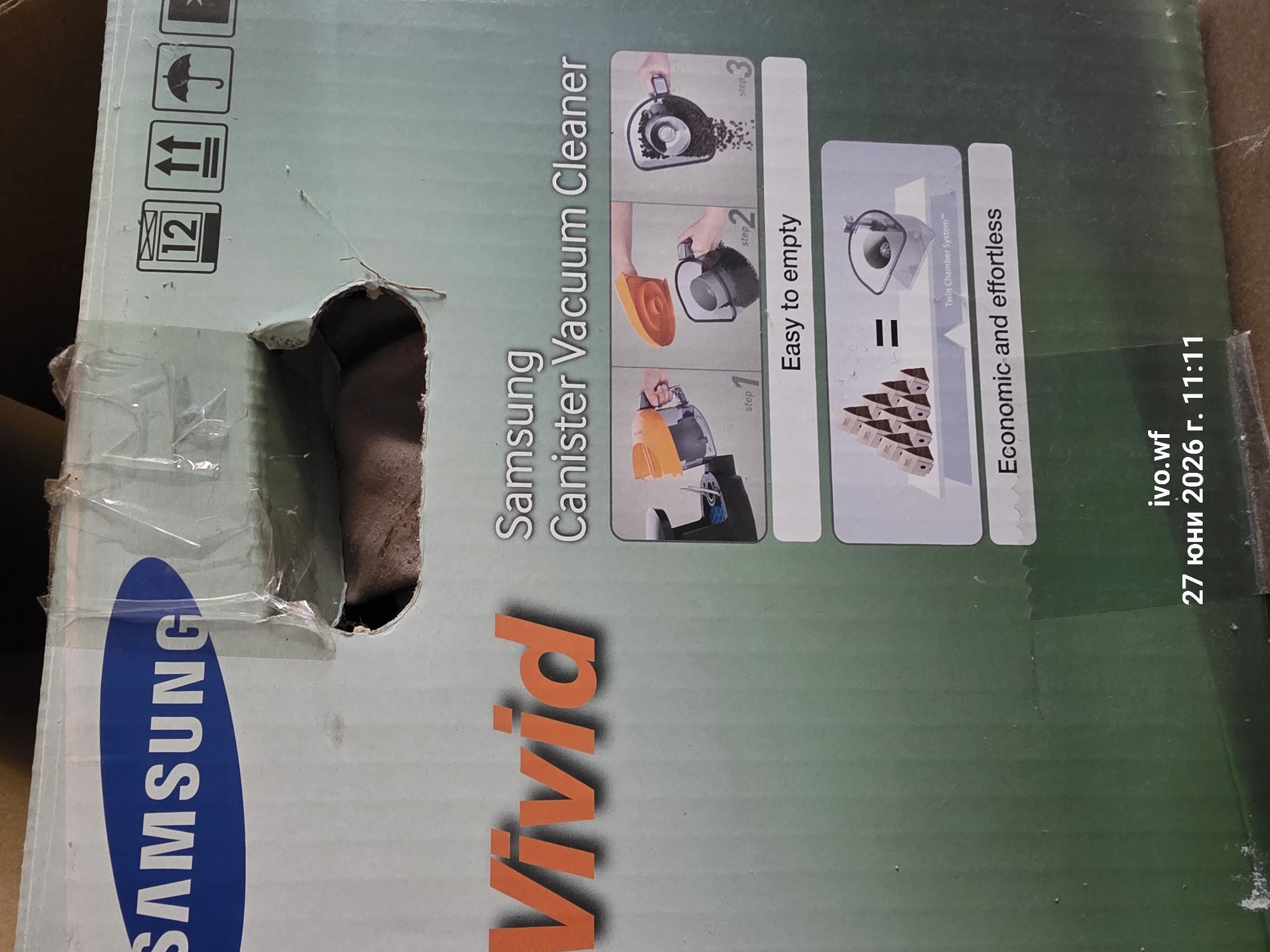 The image shows a cardboard box for a Samsung Vivid Canister Vacuum Cleaner. The box has the Samsung logo in blue and white, and the word "Vivid" in large orange letters. There are also diagrams illustrating how to empty the vacuum cleaner, labeled "Easy to empty" and "Economic and effortless." Shipping symbols are visible on the top right of the box. The box appears to be damaged in the upper left corner. The bottom of the image contains text that reads "ivo.wf 27 юни 2026 г. 11:11". The exact location within the city of Chal, Bulgaria is not provided.