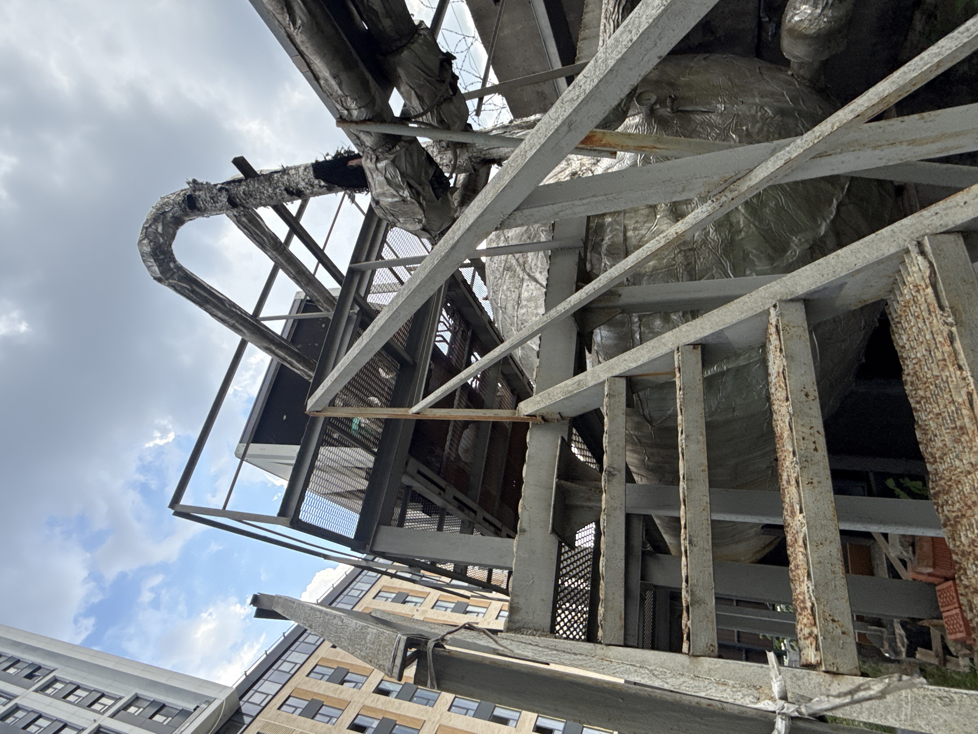The image shows a low-angle, upward view of a complex array of industrial metal scaffolding and insulated pipes against a backdrop of cloudy skies and the upper stories of urban buildings. The metal structures are visibly weathered, with some rust and corrosion. The insulated pipes are covered in foil or similar material, some of which is damaged or peeling. The composition suggests an exterior installation, possibly on the roof or side of a building in a dense urban area. There is no information in the image to confirm the location of Khimki, Russia. There are no people visible in the image.